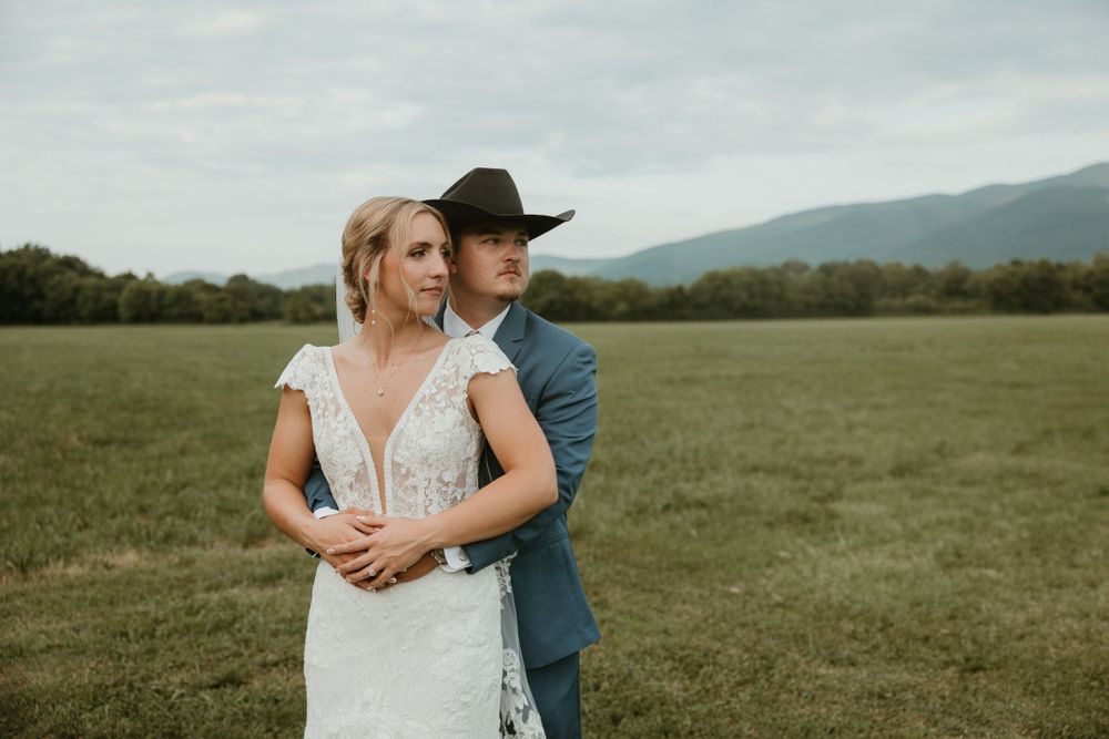 Couple embracing in rustic outdoor wedding setting with rolling hills in background.