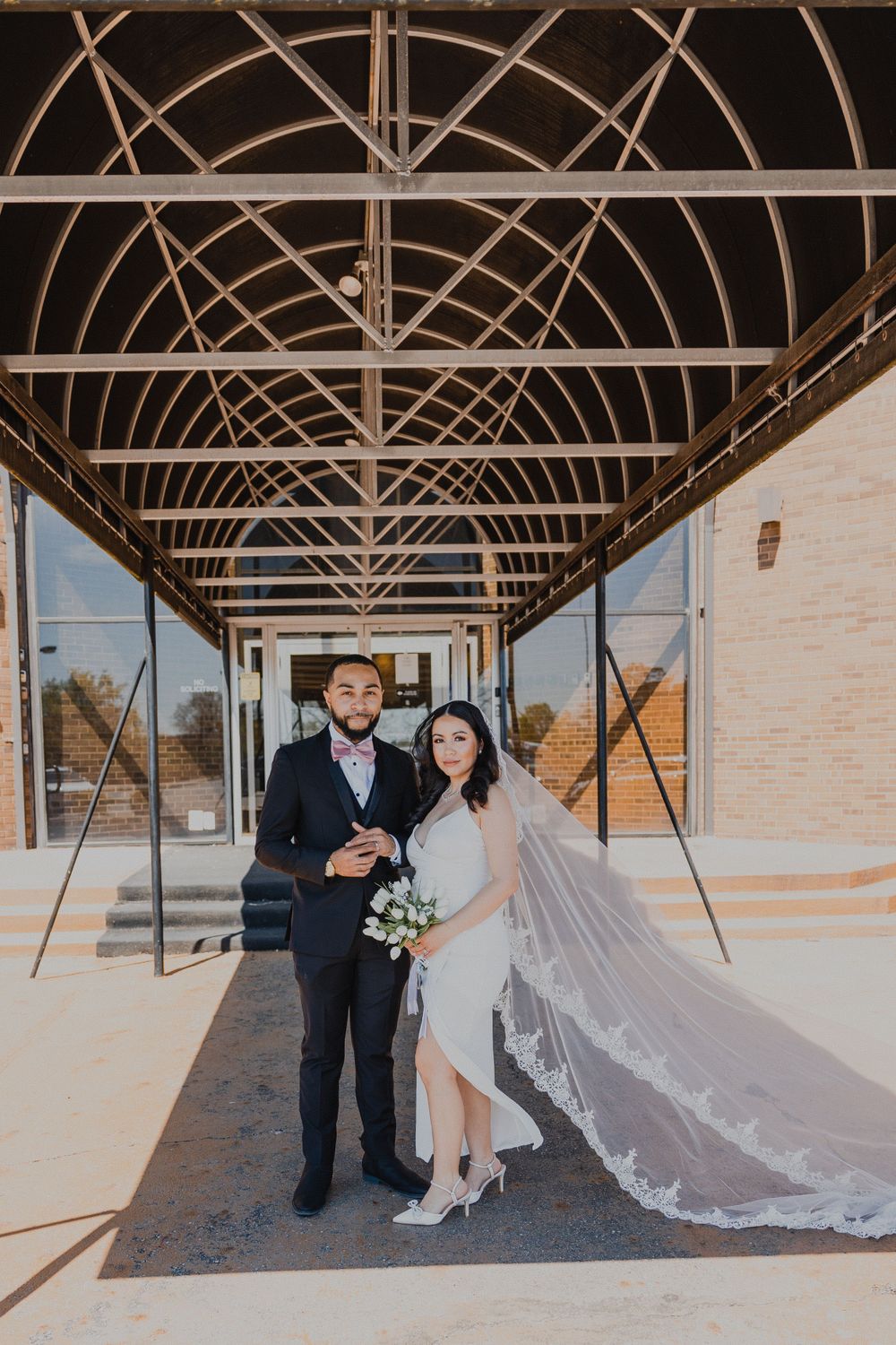 Wedding couple poses under architectural beams in covered walkway with long flowing veil and formal attire.