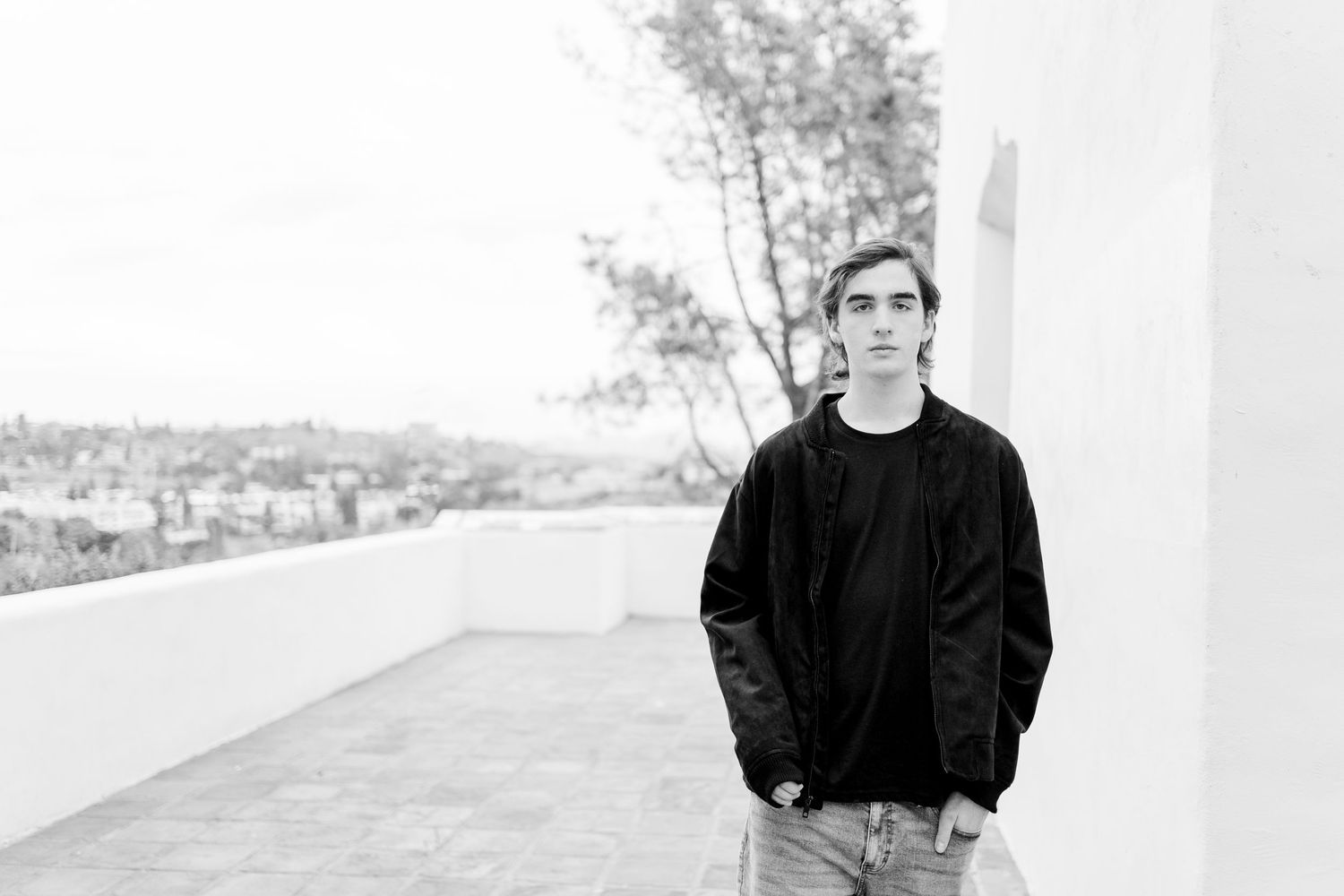 A black and white portrait of someone in casual attire standing on a balcony overlooking a distant cityscape.