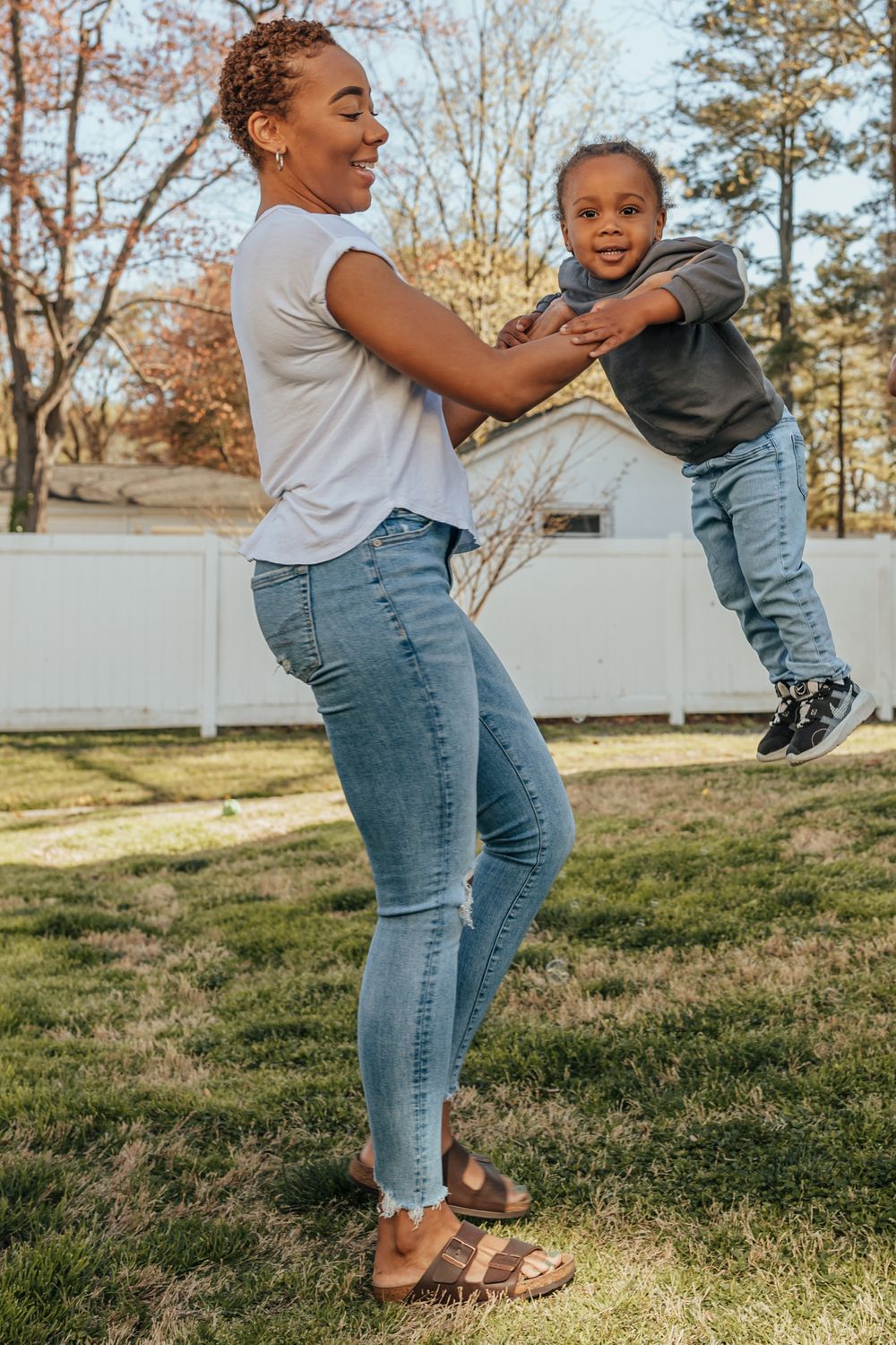 Parent and child share a playful moment swinging and laughing together in a backyard with white fence on sunny day.
