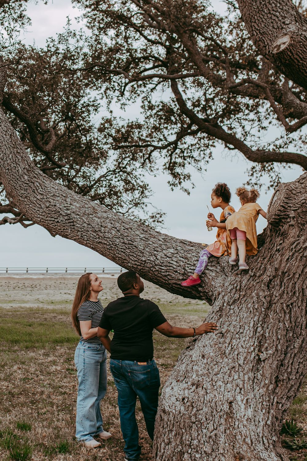 A large oak tree frames a rustic outdoor family scene with two children sitting on a low branch and adults standing below.