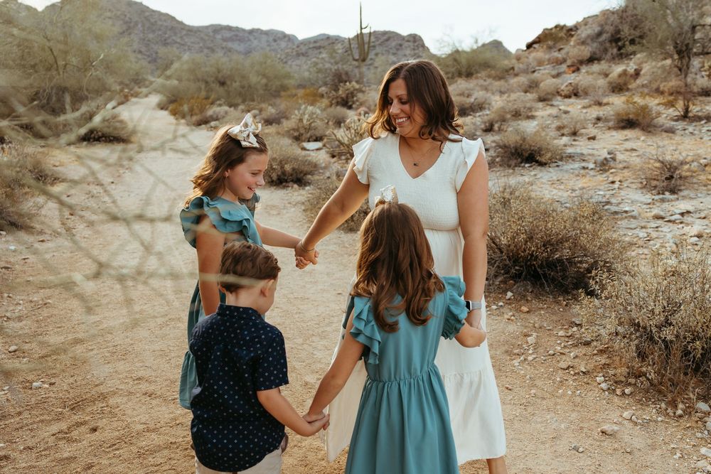 A mother holds hands with her three children in a desert landscape with cacti and mountains in the background.