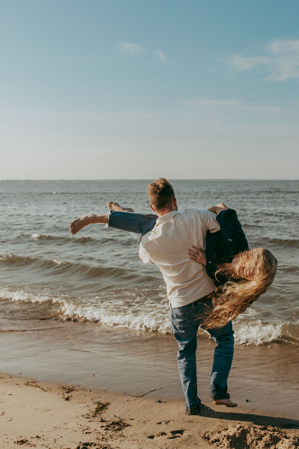 A couple dance playfully together on the beach at sunset with waves rolling in along the shoreline.