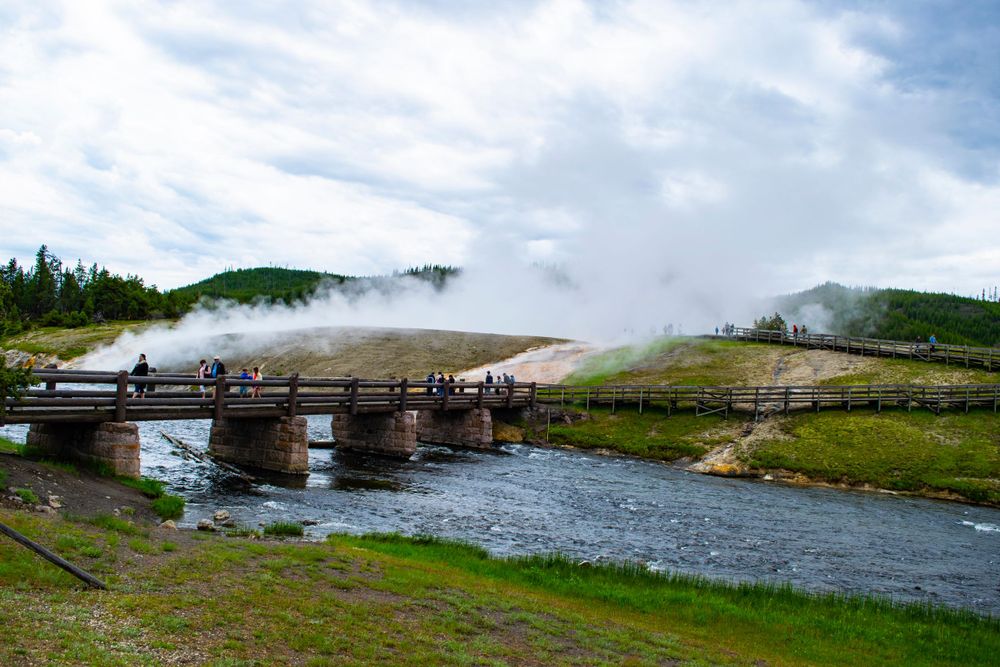 Wooden footbridge crosses river with steam vents and geothermal features in mountainous landscape.