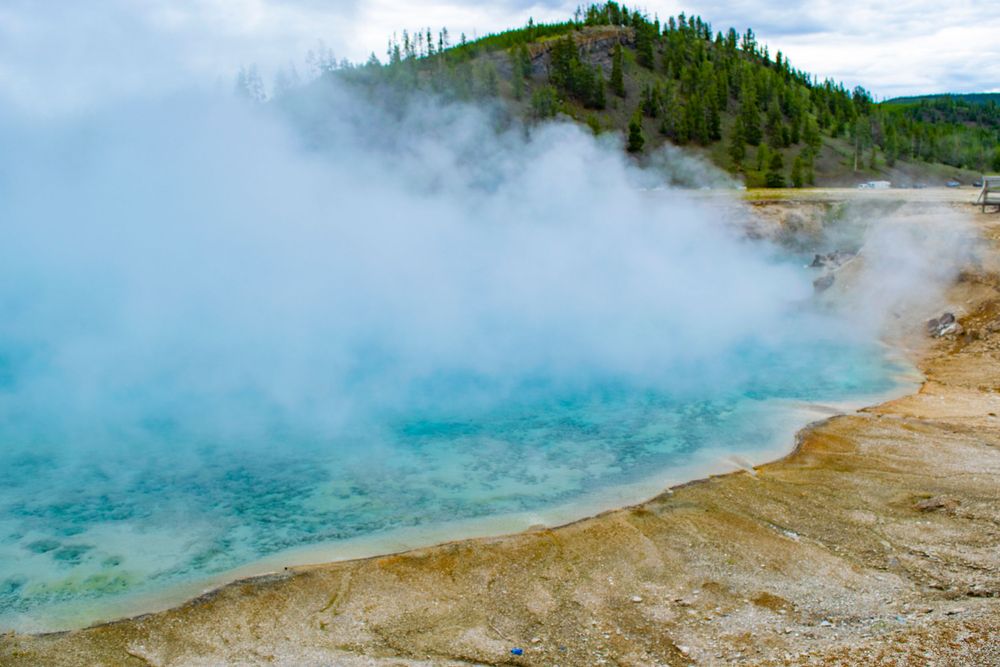Steaming turquoise hot spring pool surrounded by mineral deposits and forested hills in Yellowstone.