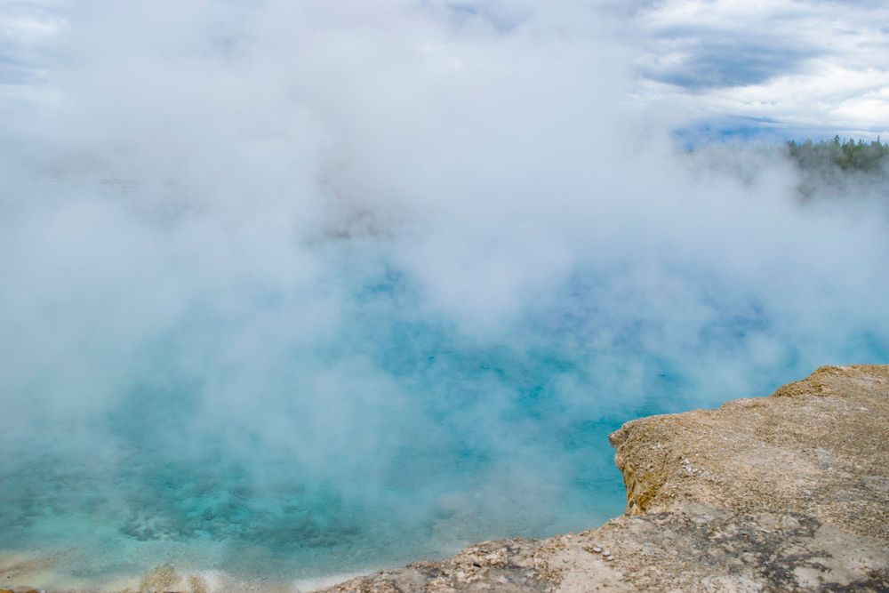 Misty blue hot spring with mineral-crusted edges against cloudy mountain backdrop.