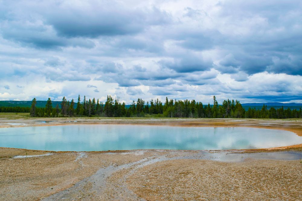Serene turquoise hot spring pool reflects cloudy sky with pine forest in background.