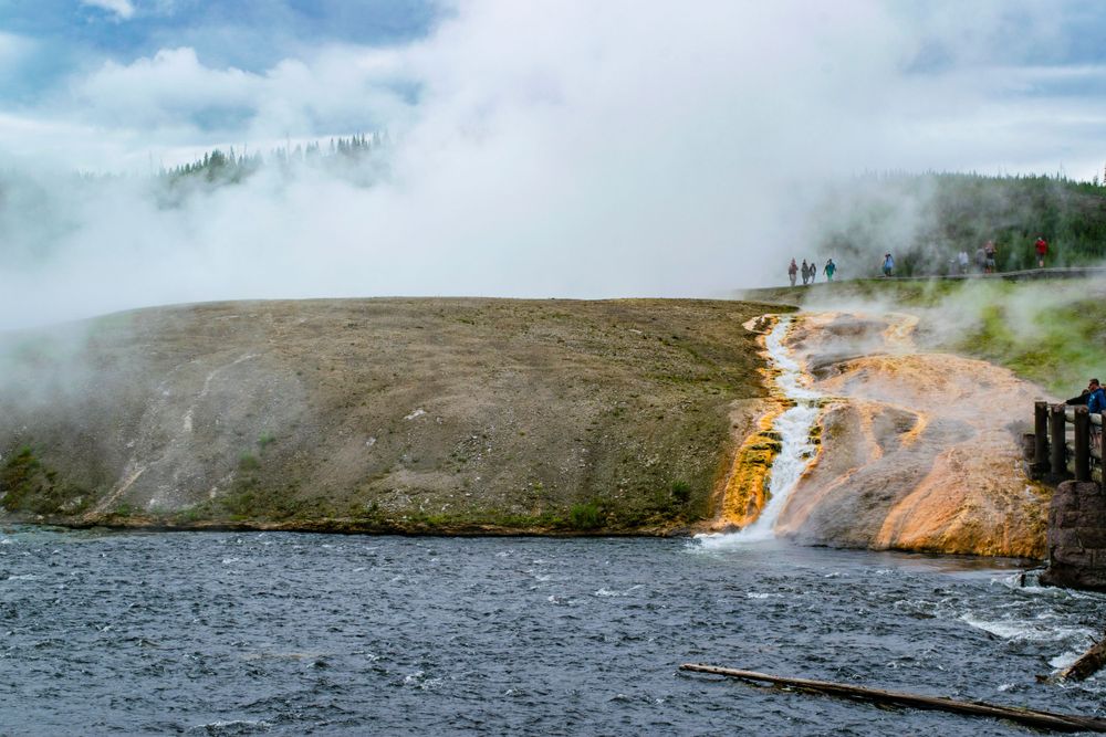 Steaming waterfall cascades down rocky cliff face into dark lake below.