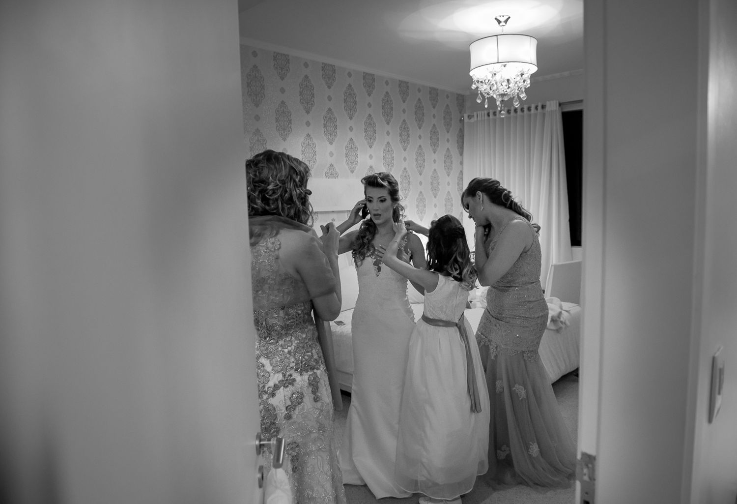 A group gathers in a room with floral wallpaper and crystal chandelier during wedding preparations.
