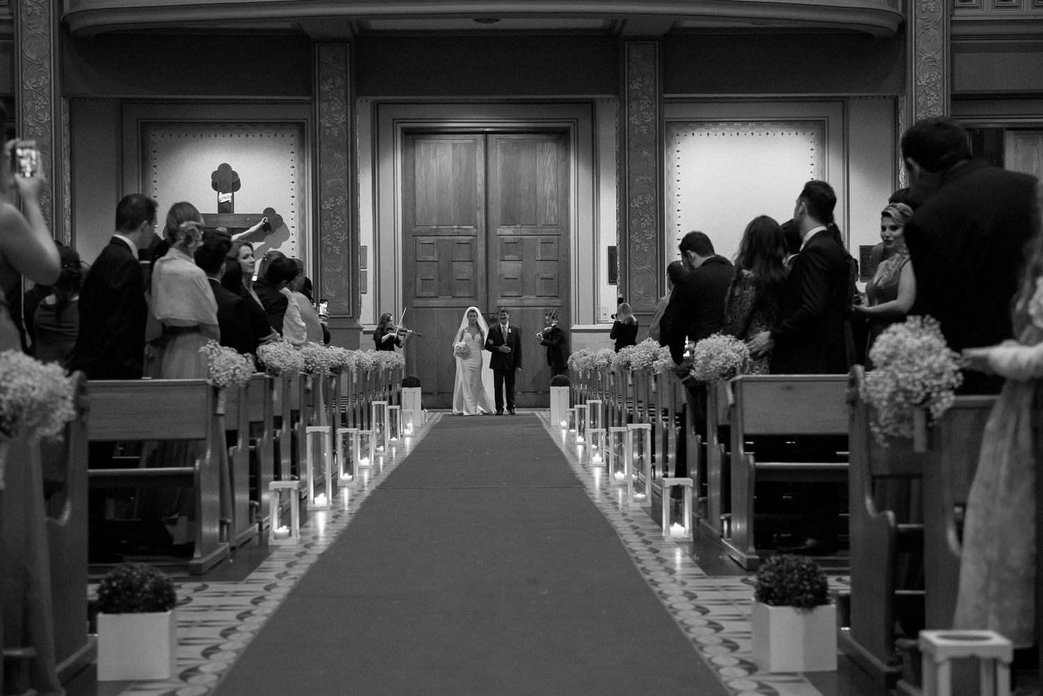 Black and white view down church aisle with guests seated along floral-decorated pews.
