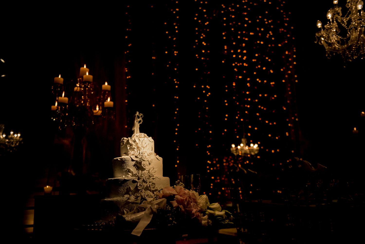 Wedding cake illuminated by candlelight and string lights in a dark reception setting.