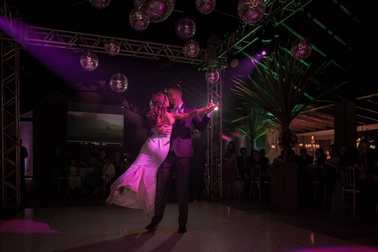A couple performs their first dance under purple lighting on a wedding reception dance floor.