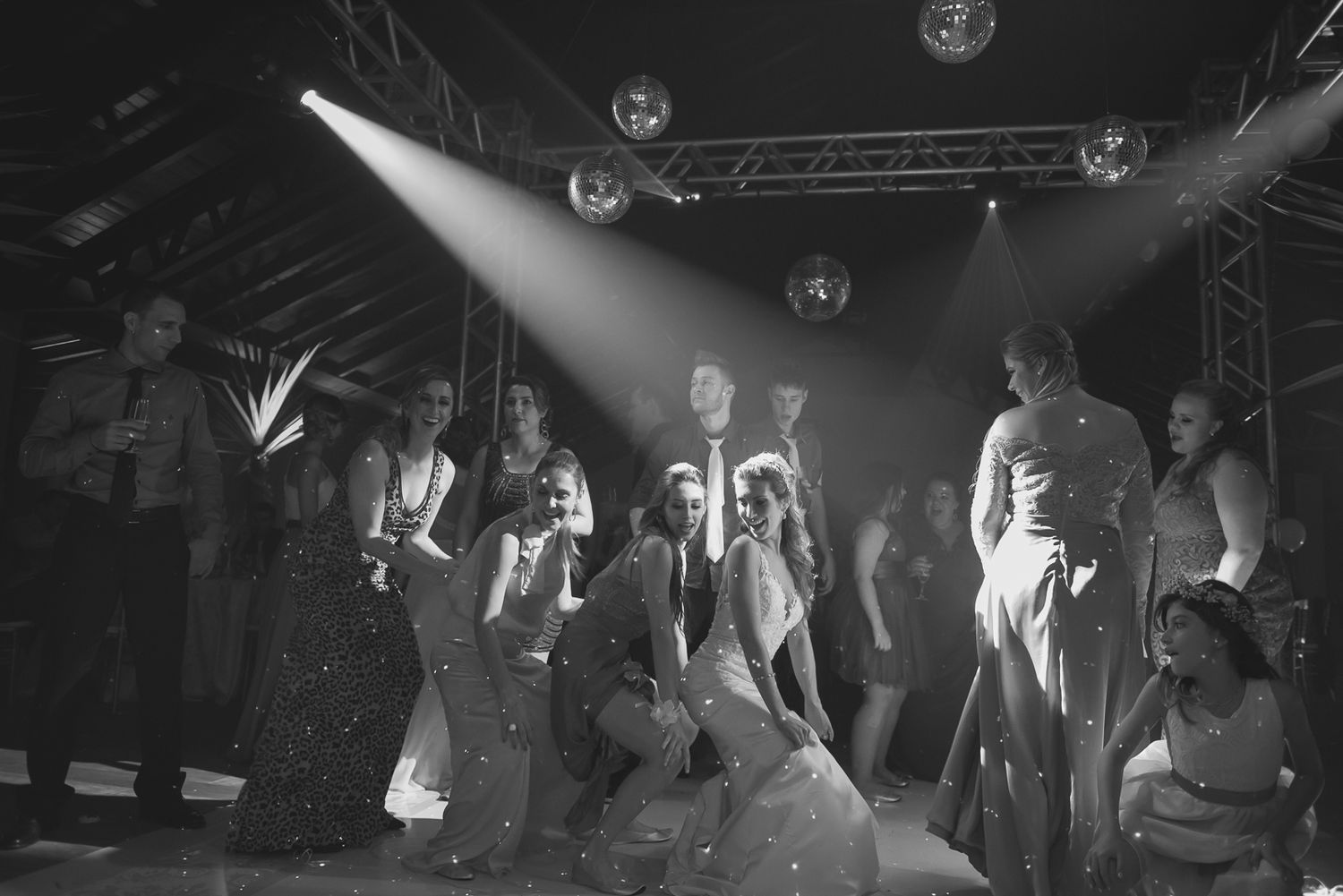 A group of wedding guests dancing under dramatic spotlights in a dark reception venue.