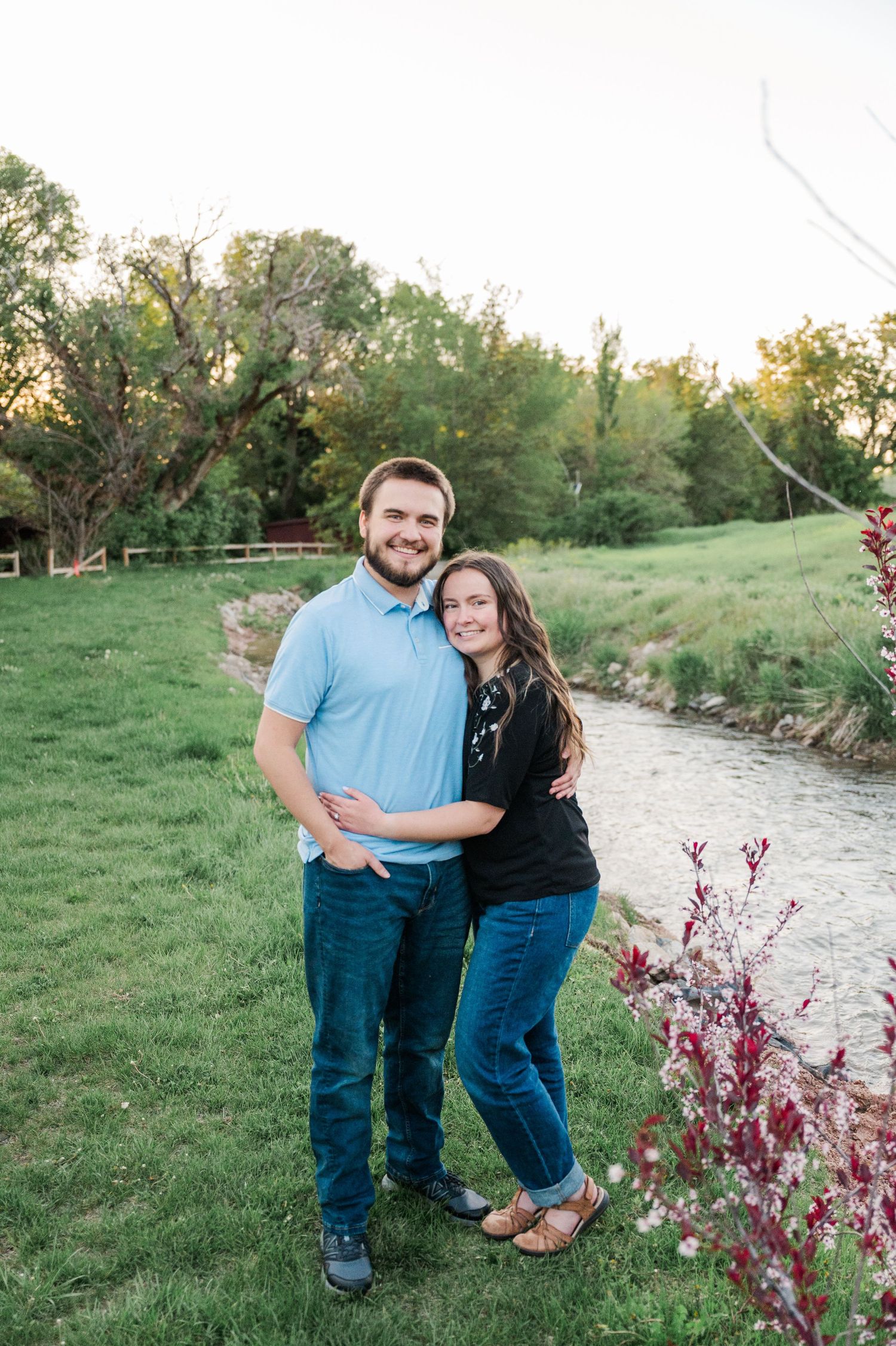 A couple poses together by a winding creek with flowering bushes and green trees in the background.