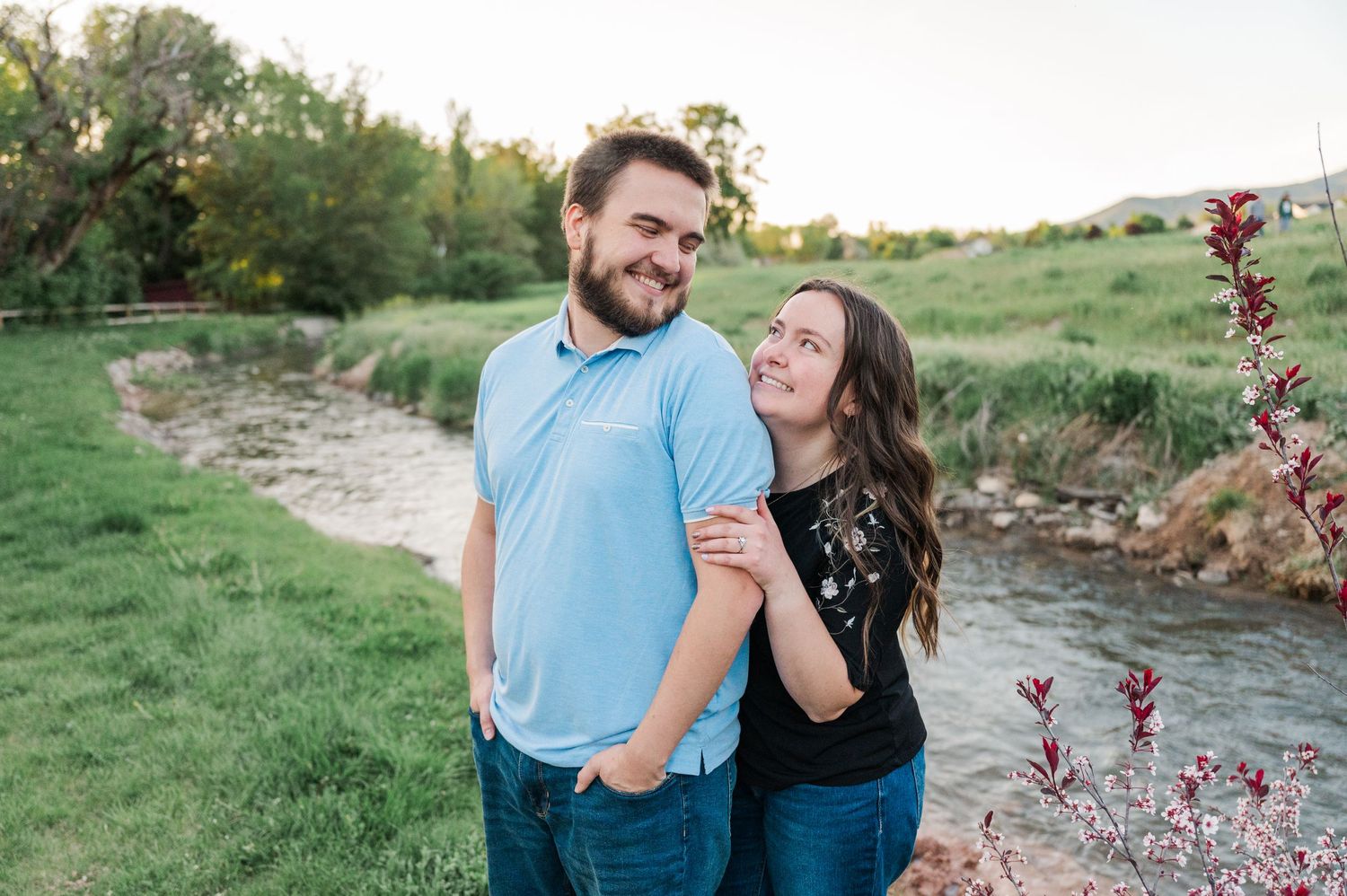 A couple shares a romantic moment while standing on grassy riverbank with spring foliage.