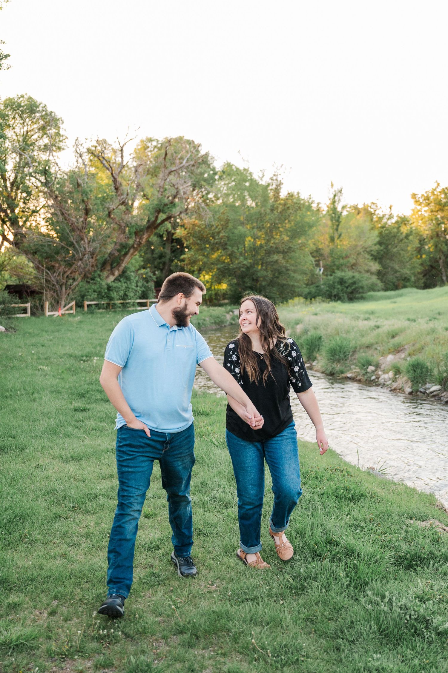 A couple walks hand in hand along a creek bank surrounded by lush green grass and trees at sunset.