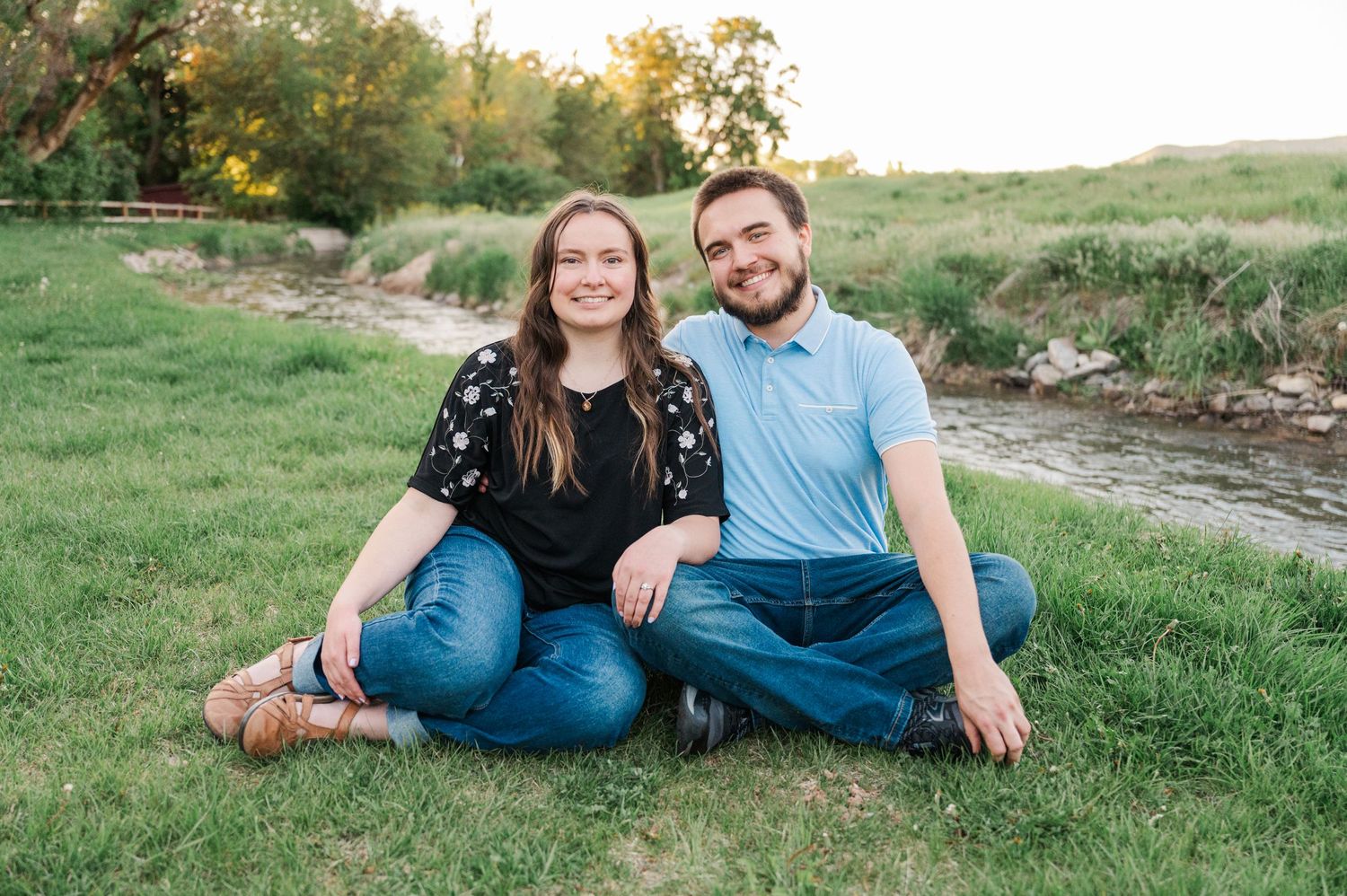 A couple sits casually on grass near a meandering creek on a sunny afternoon.