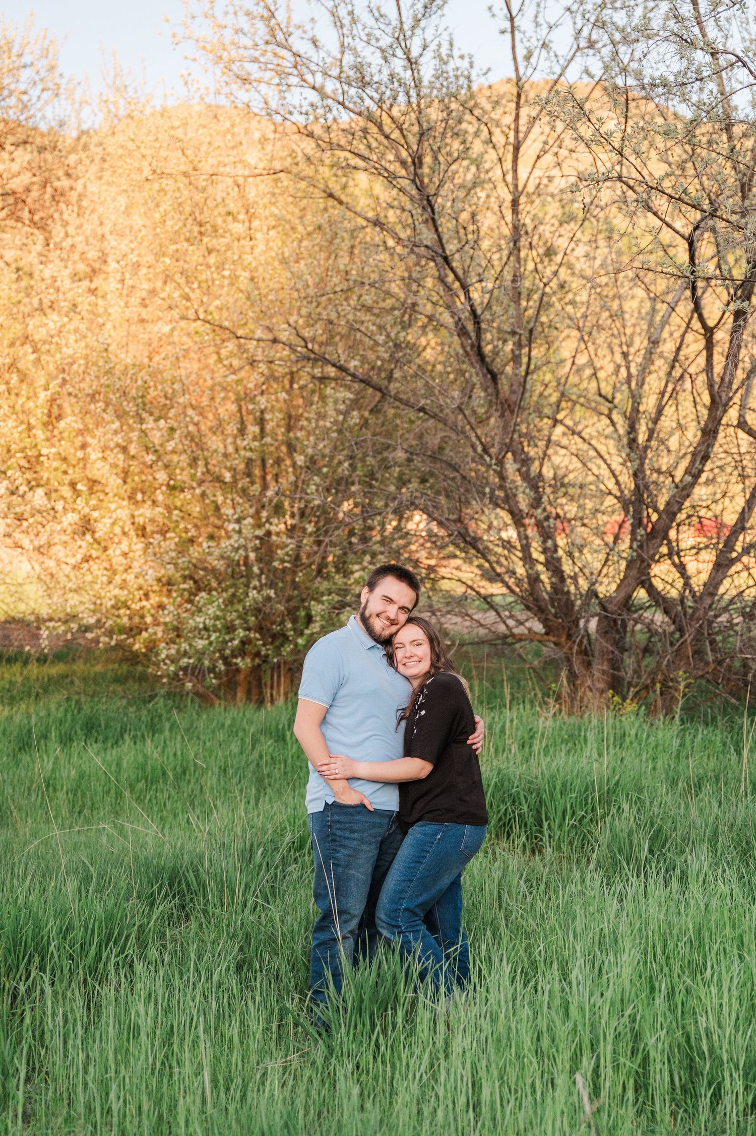 A couple embraces in tall grass with golden autumn trees glowing in the background.