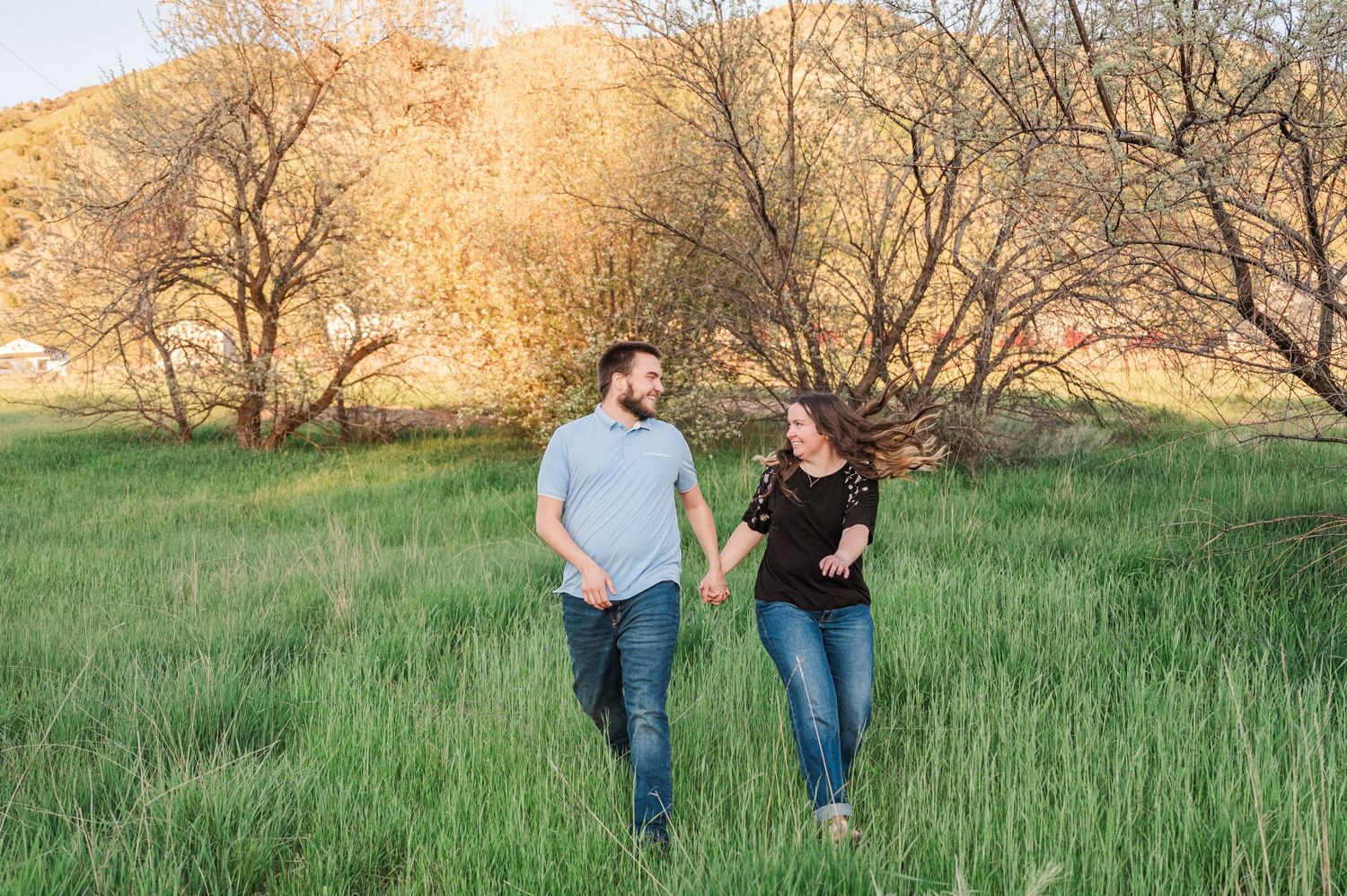 A couple walks together through a field of tall grass with golden sunlit trees behind them.