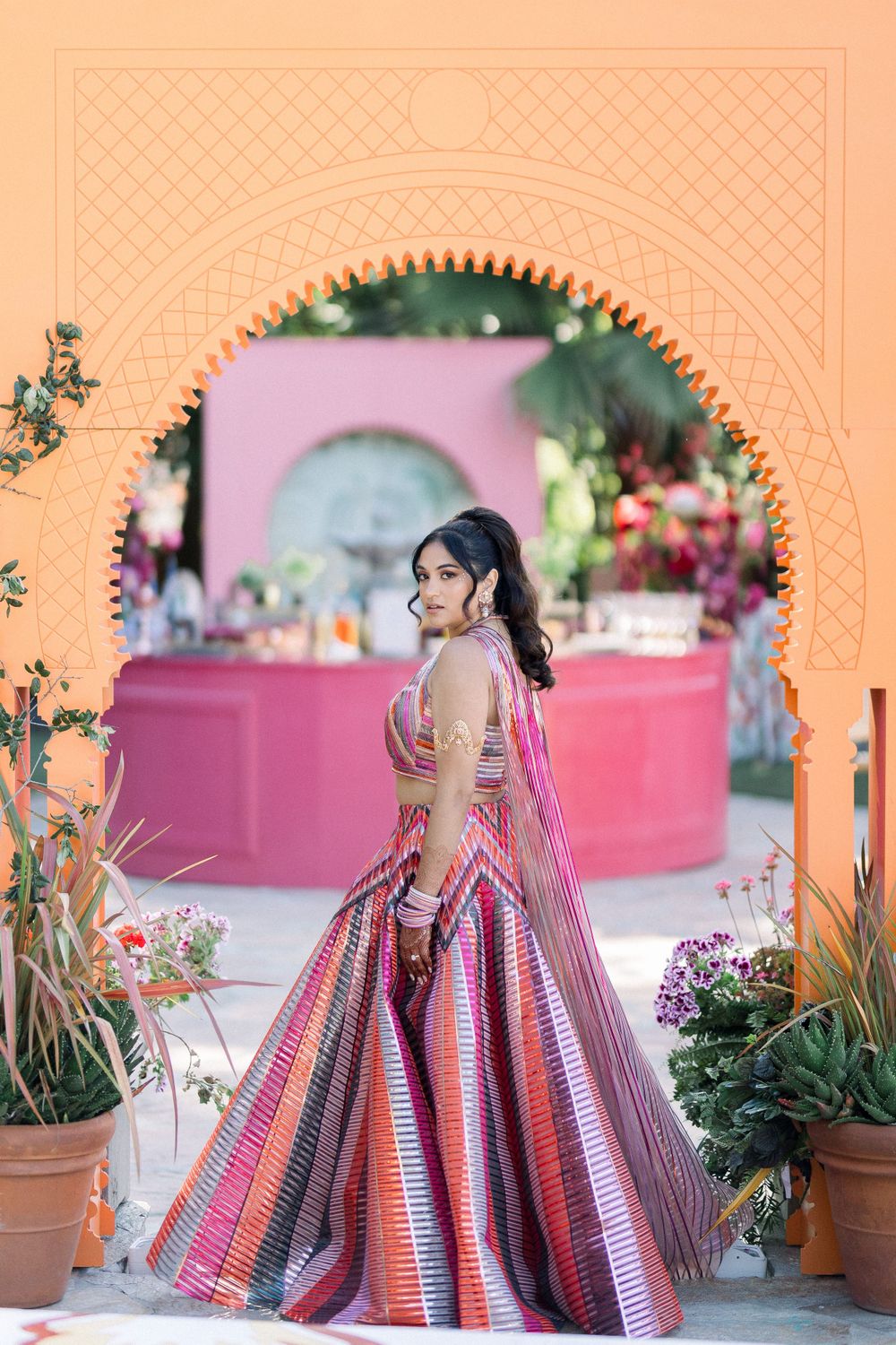 Ornate wedding lehenga in vibrant pink and red tones photographed against a coral-colored archway.