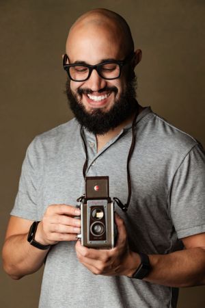 Enthusiastic photographer holding vintage twin lens reflex camera while wearing glasses and grey t-shirt against brown backdrop.