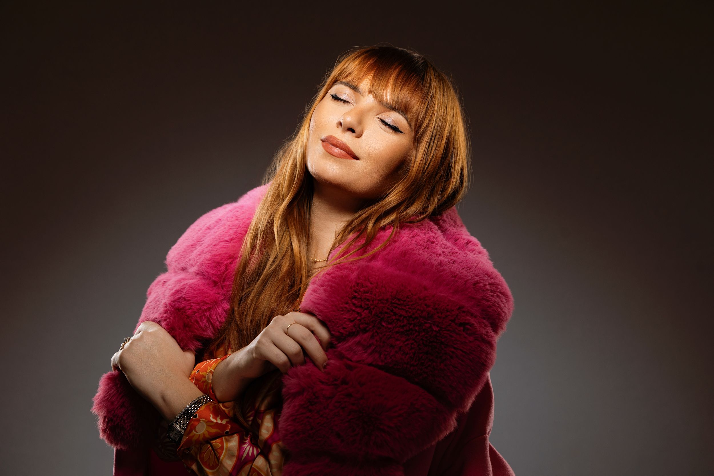 A figure in a luxurious fuchsia fur coat poses with eyes closed against a dark backdrop.