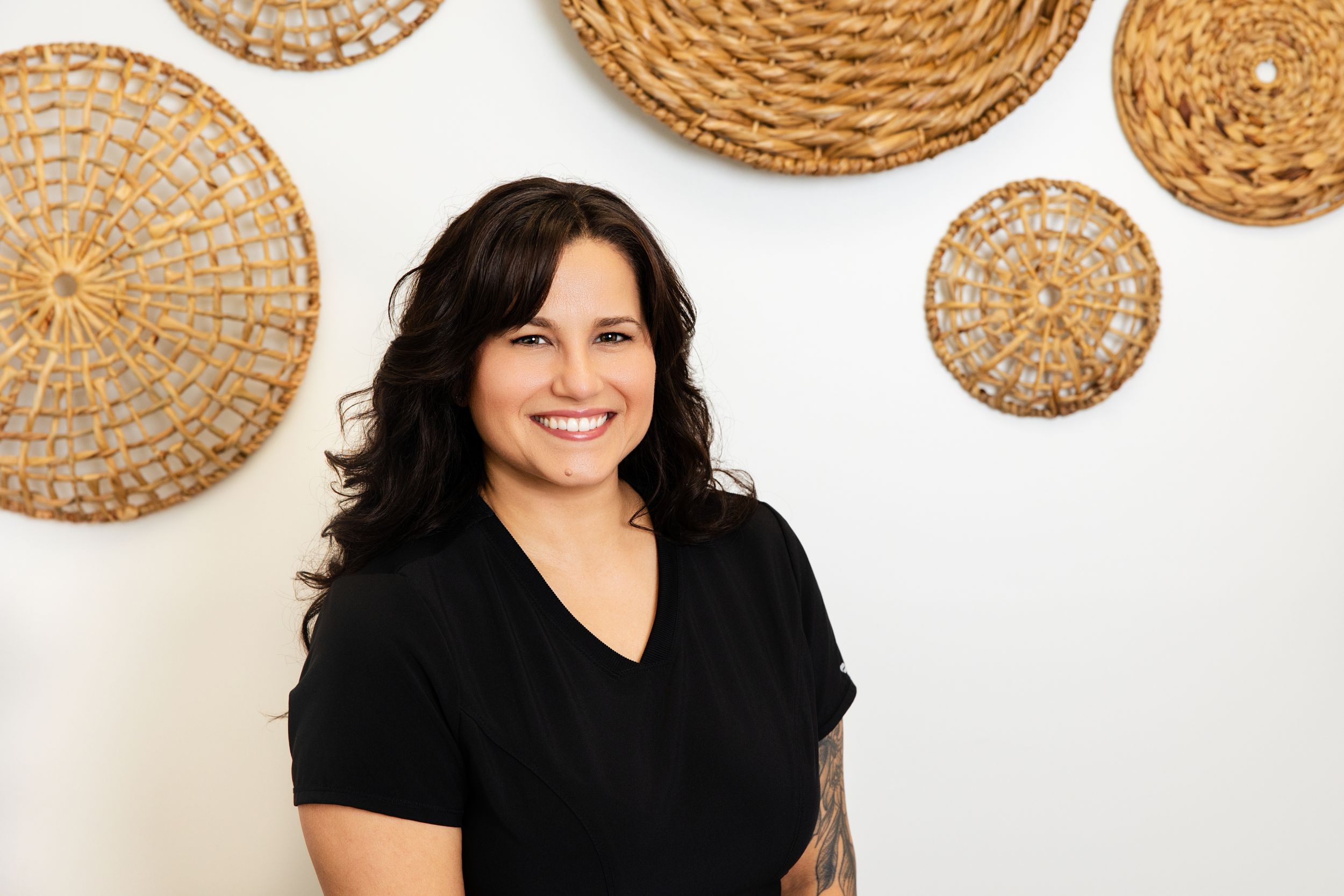 Woman in black top smiles while standing against white wall decorated with woven wicker baskets.