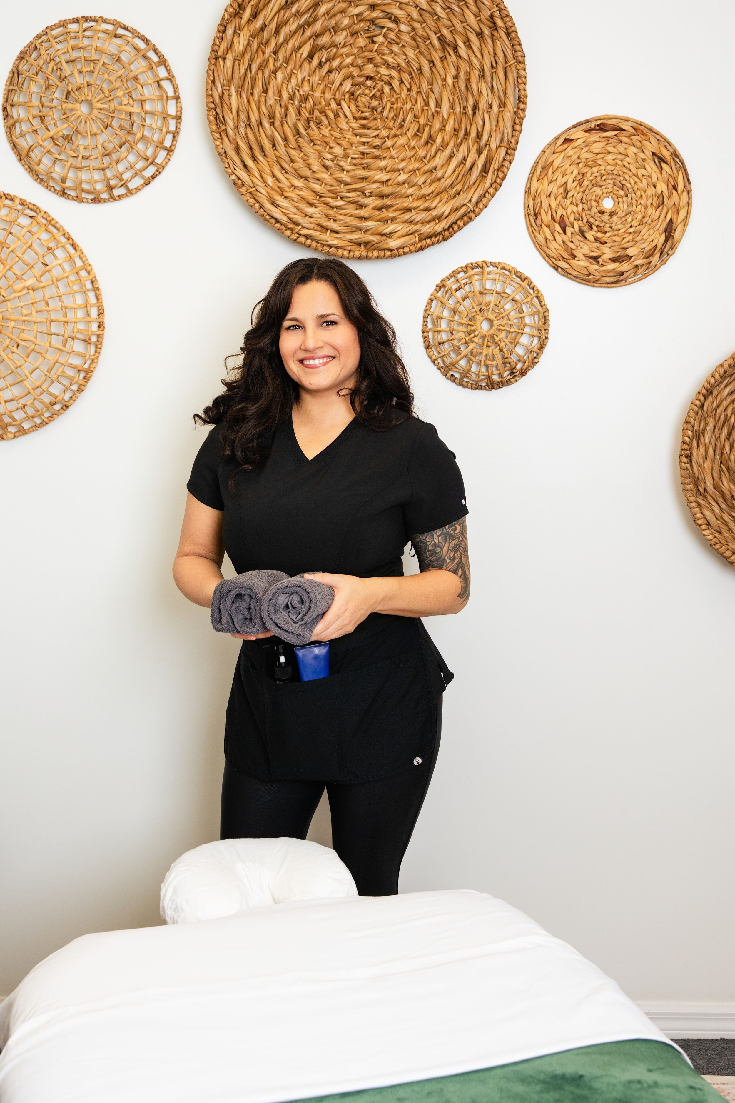 Professional massage therapist standing in treatment room with decorative woven baskets on white wall.