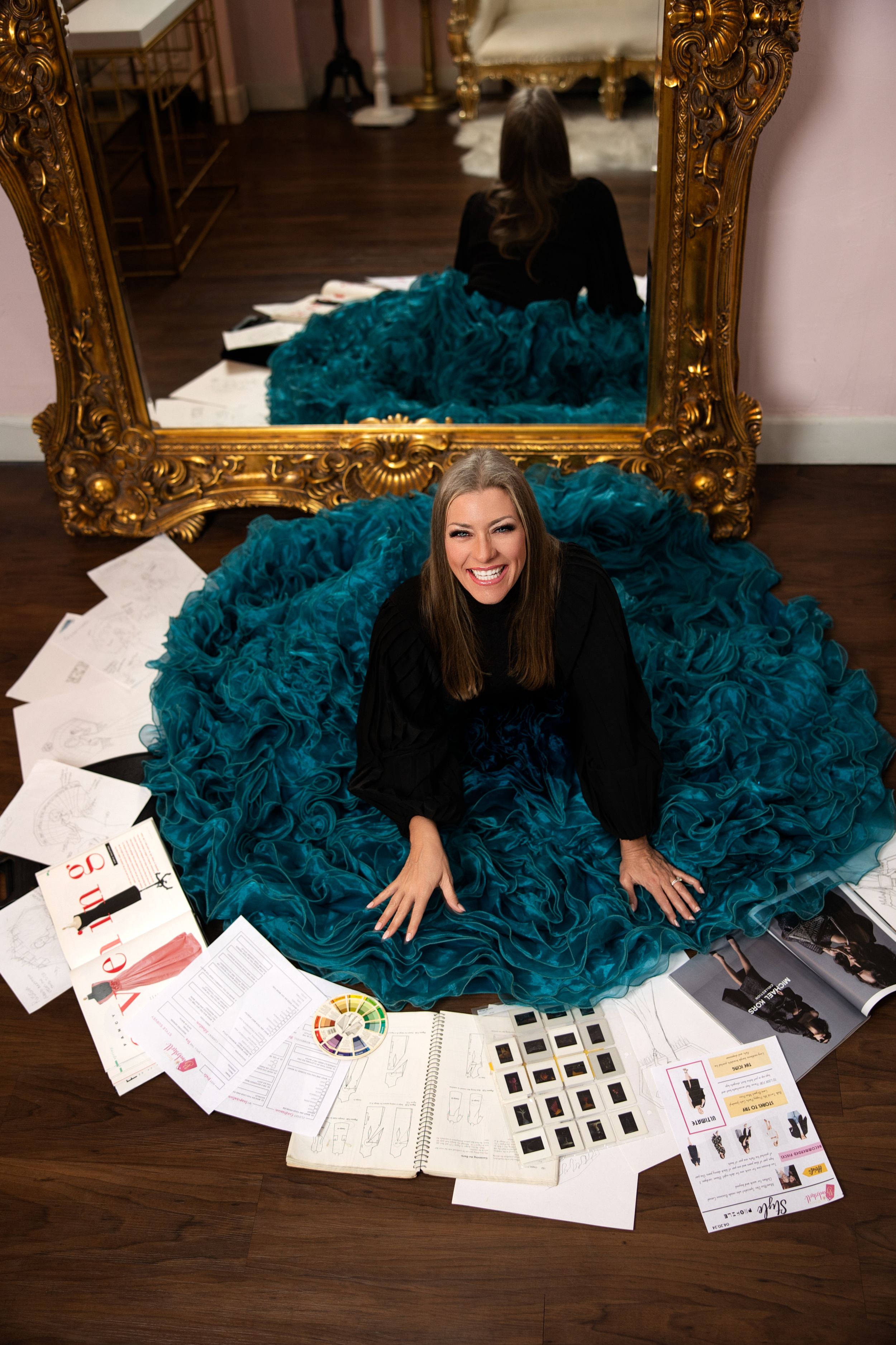 Woman in black sweater sits on teal tulle skirt surrounded by design sketches and papers on hardwood floor with gold mirror.