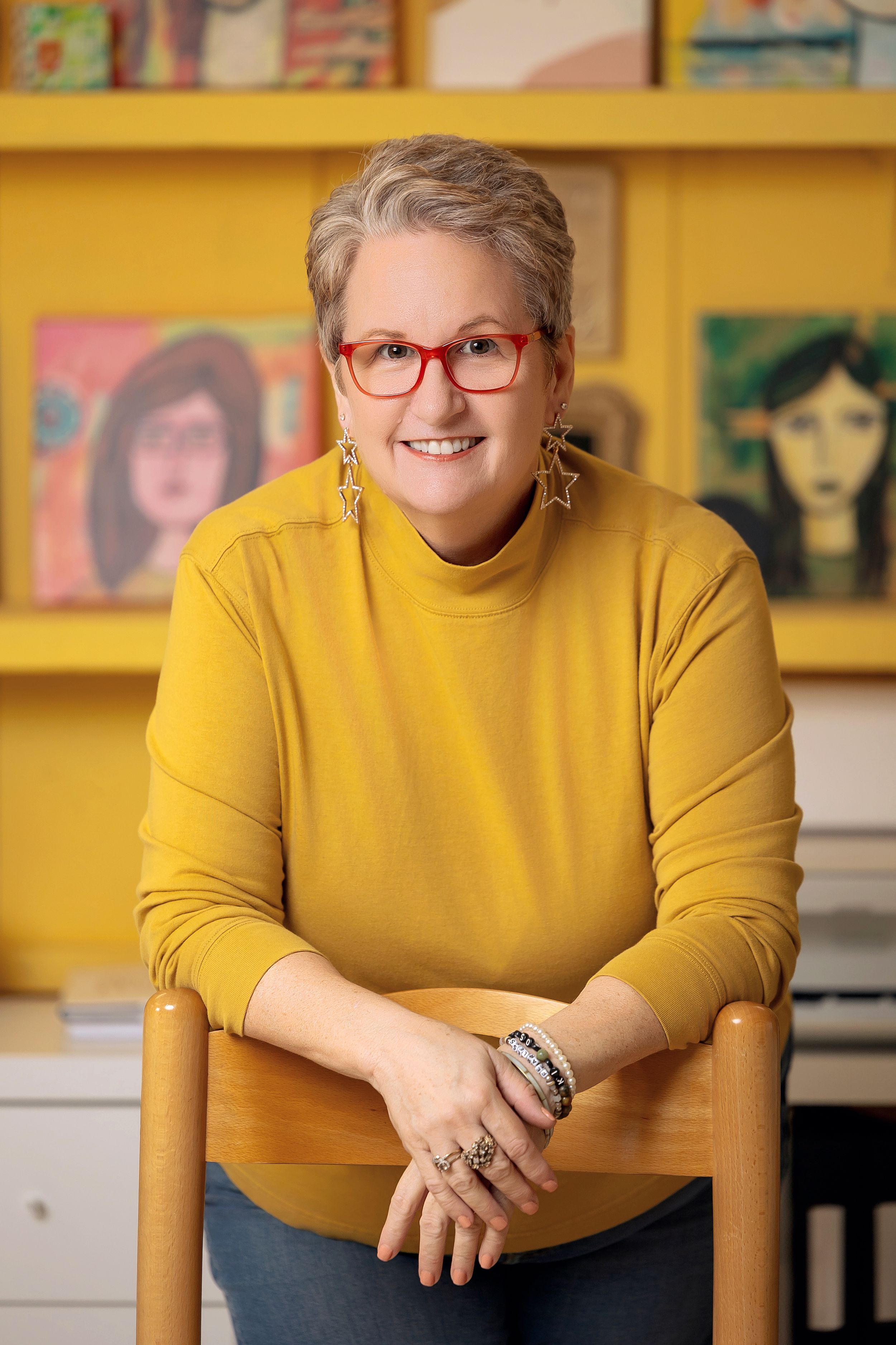 Person in yellow sweater and red glasses sits in wooden chair against backdrop of colorful artwork on yellow shelves.