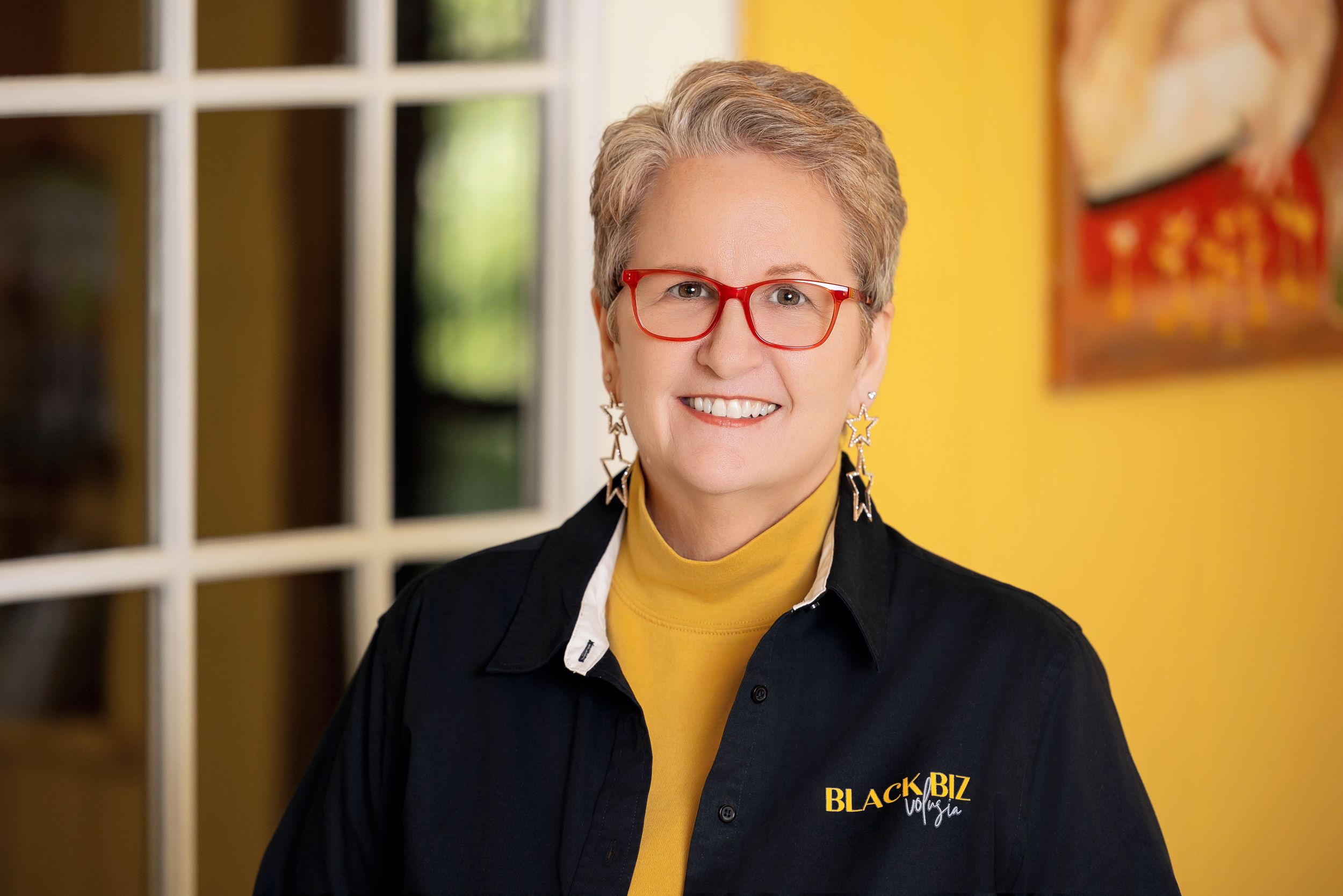 Professional portrait of someone wearing a yellow turtleneck sweater and navy blazer with BR Media logo against a blurred interior.