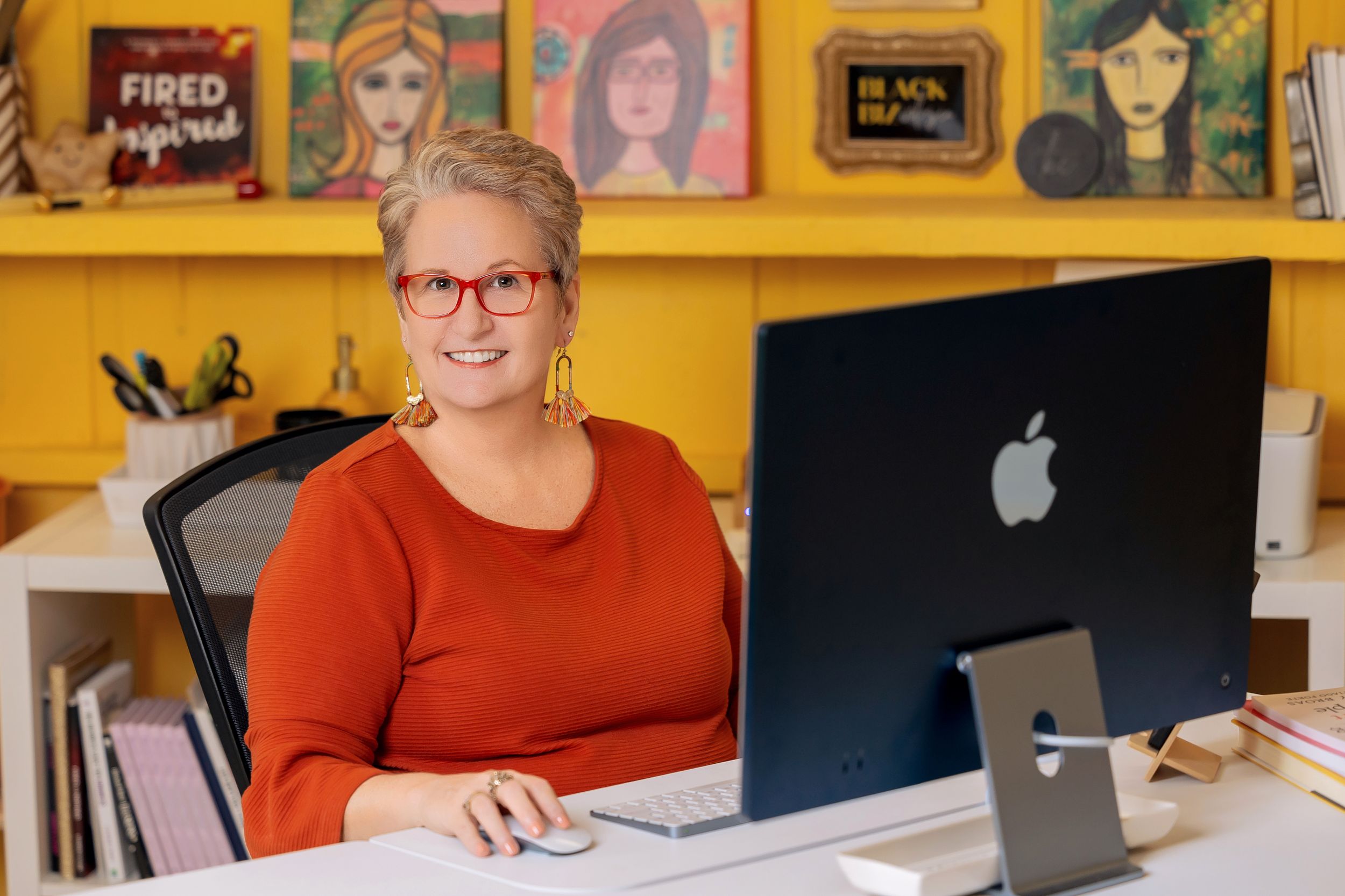 A person in a red sweater works at a desk with Apple computers and colorful album artwork displayed on shelves behind them.