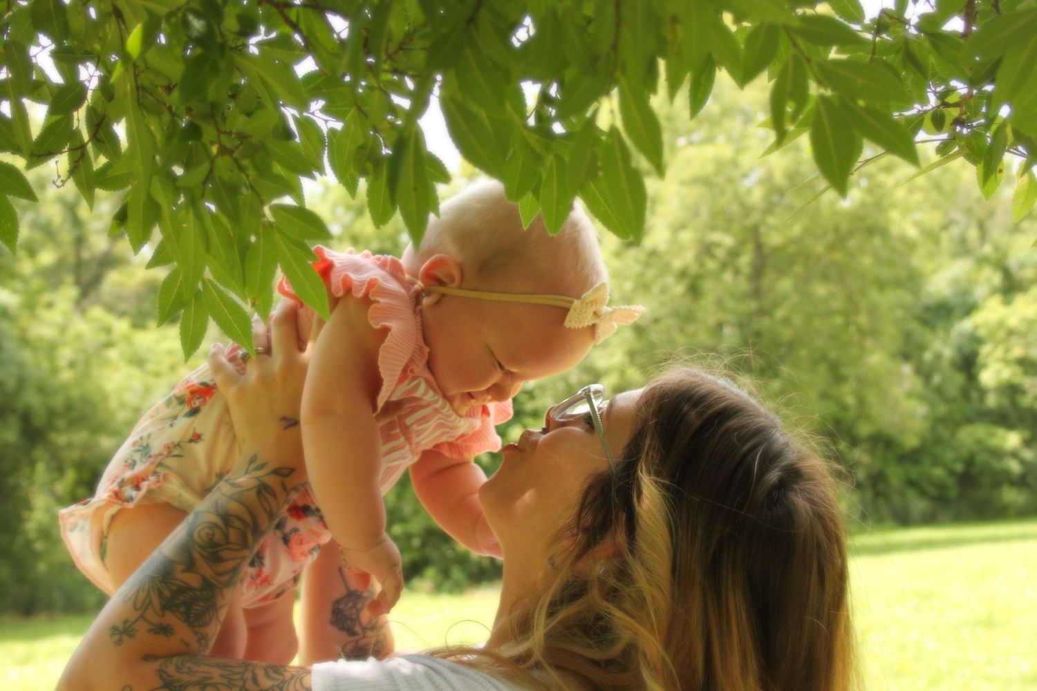 Mom holding up her baby daughter kissing her in front of trees.
