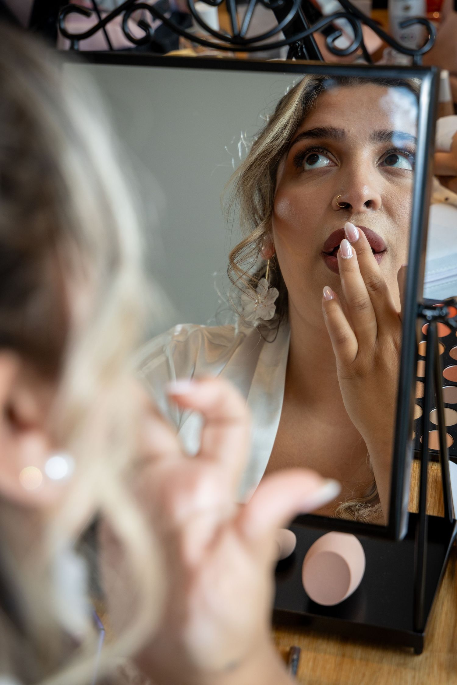 Bride applying lipstick while gazing into an ornate mirror during wedding day preparations.