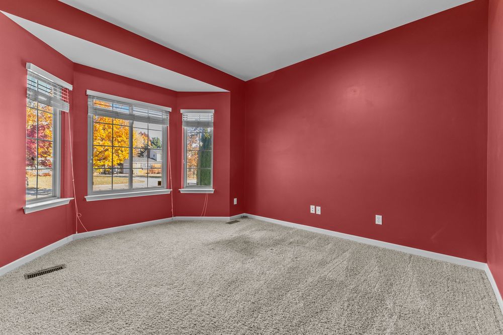 Bright bedroom with bold red accent walls, carpet flooring, and large bay window with natural light.