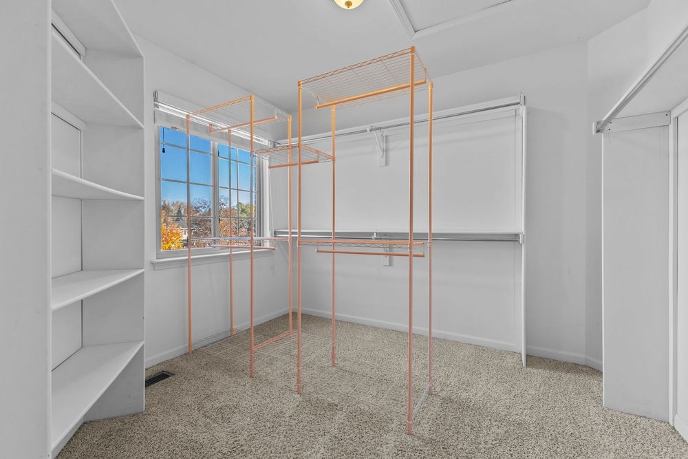 Spacious walk-in closet with white shelving, orange metal frame organizers, and carpeted floors with a bright window.