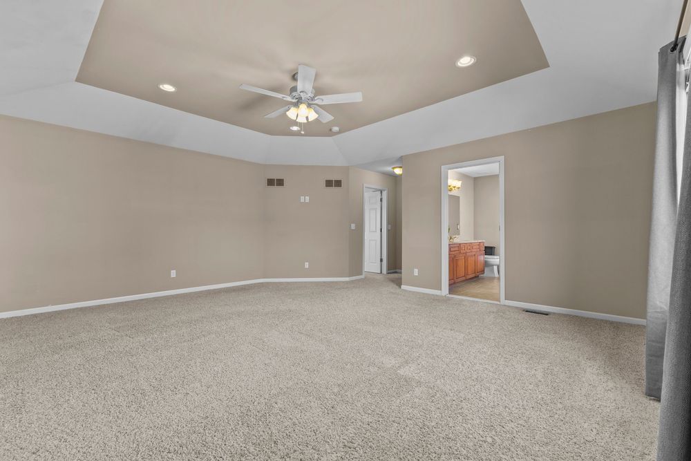 Large carpeted bedroom with tray ceiling, ceiling fan, and neutral beige walls with an open doorway.