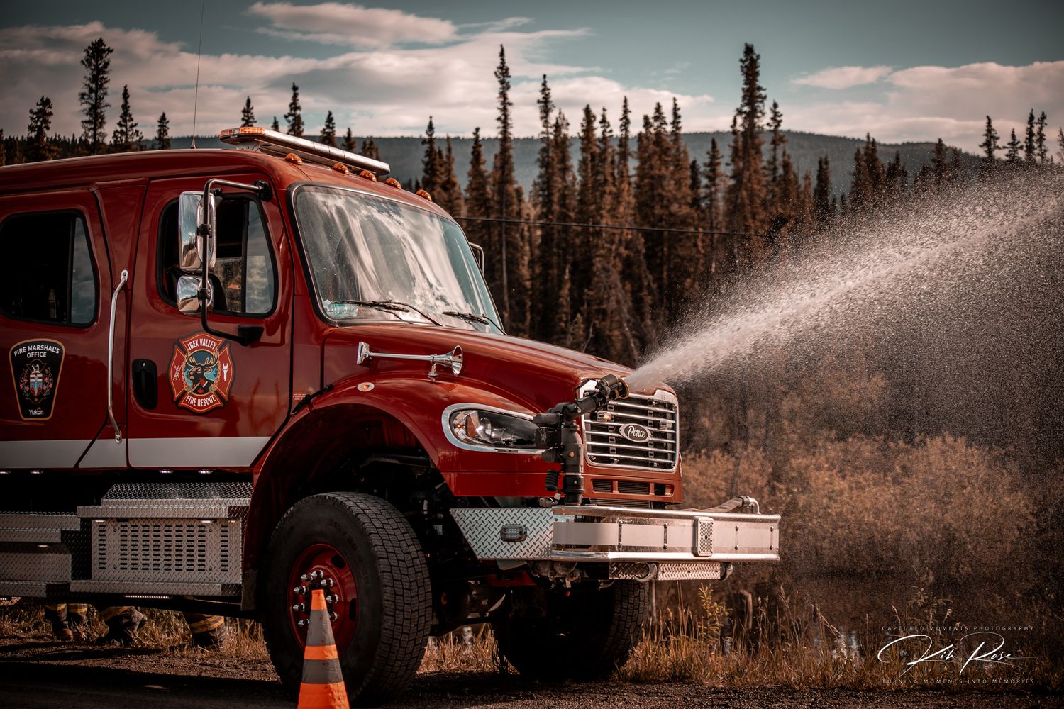 Red fire truck spraying water on a rural highway surrounded by autumn trees.