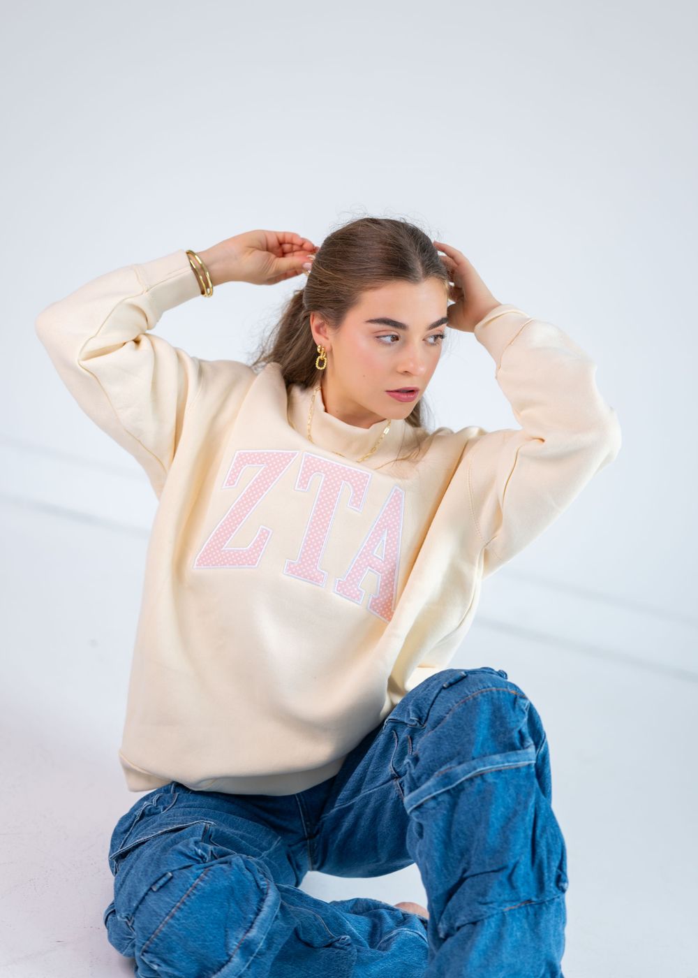 Young woman sitting on floor wearing cream ZTB graphic sweatshirt with pink letters and blue jeans.