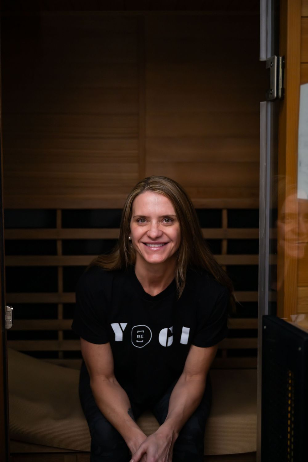 Smiling woman in black YOGA t-shirt sitting in a wooden infrared sauna, radiating wellness and relaxation.
