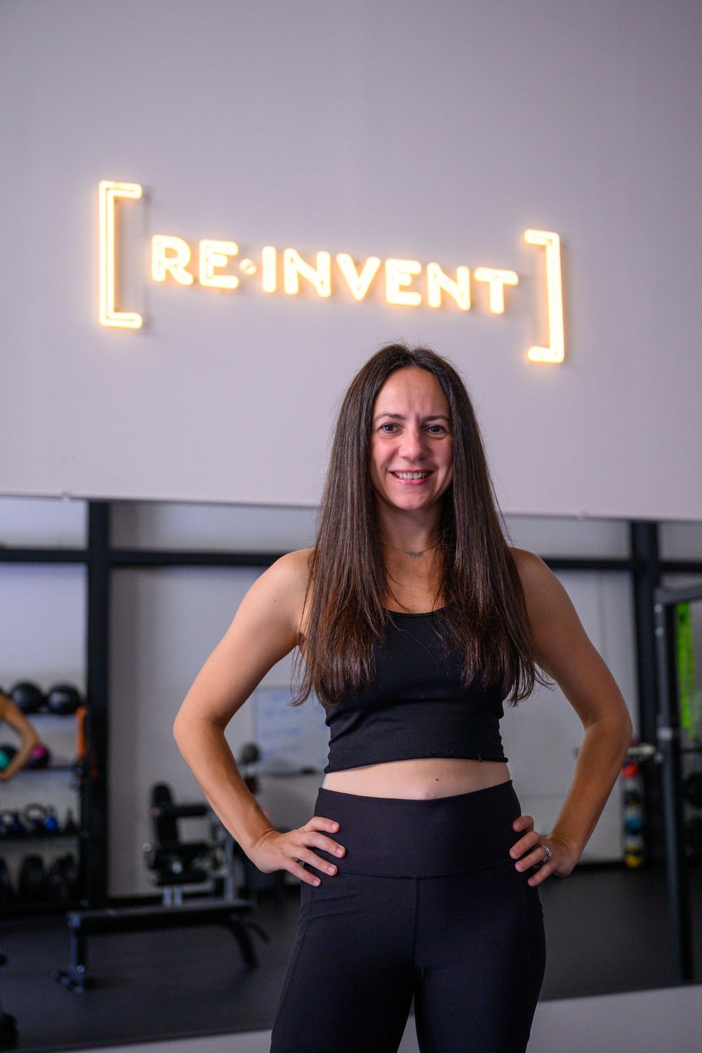 A person in black workout attire poses confidently in a fitness studio with a 'Reinvent' neon sign on the wall.