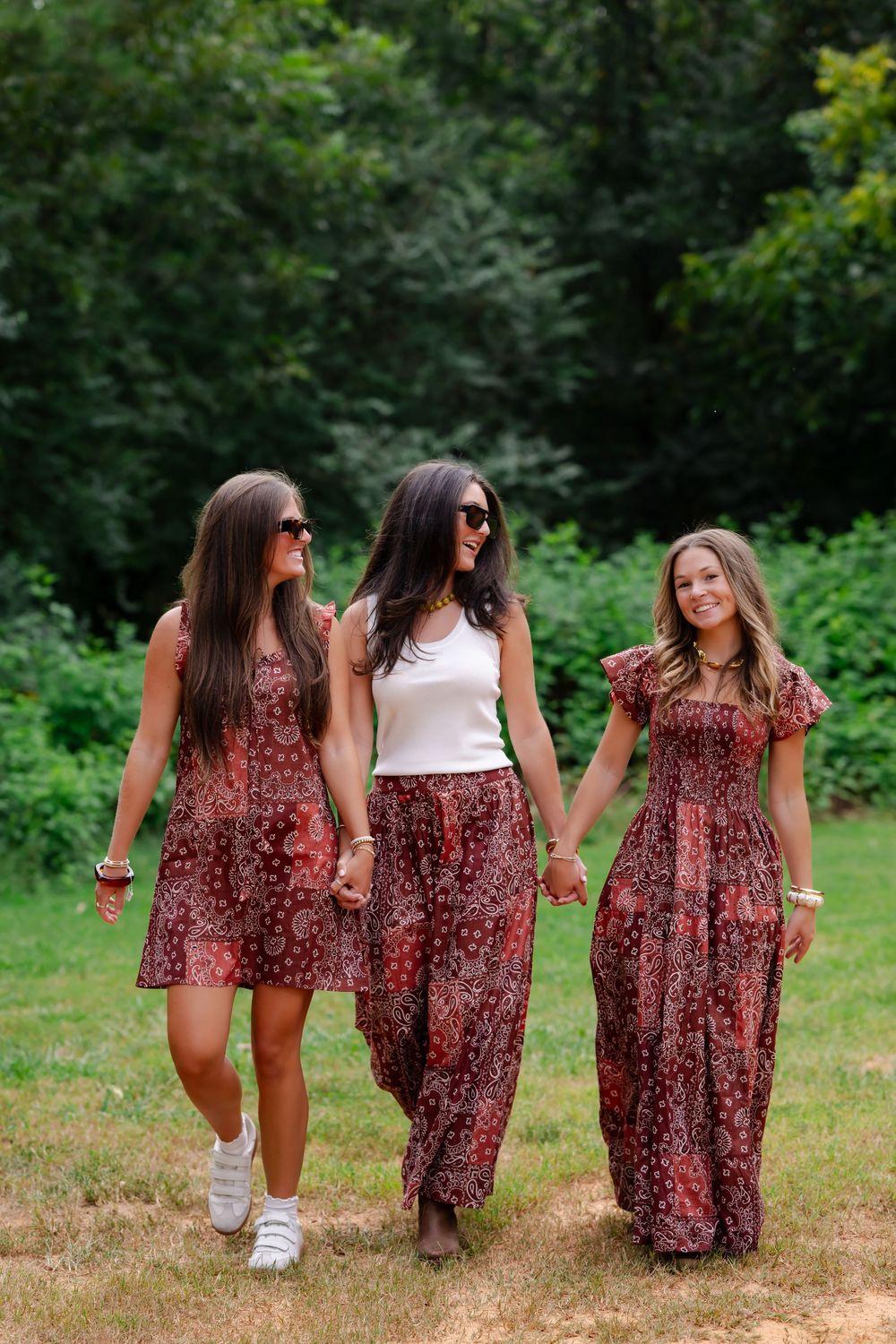 Three women in matching red floral outfits hold hands walking outdoors in a lush green park setting.