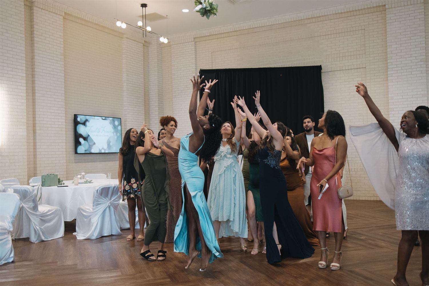 A group of guests in formal attire reach up to catch a bouquet at an elegant wedding reception in a bright ballroom.