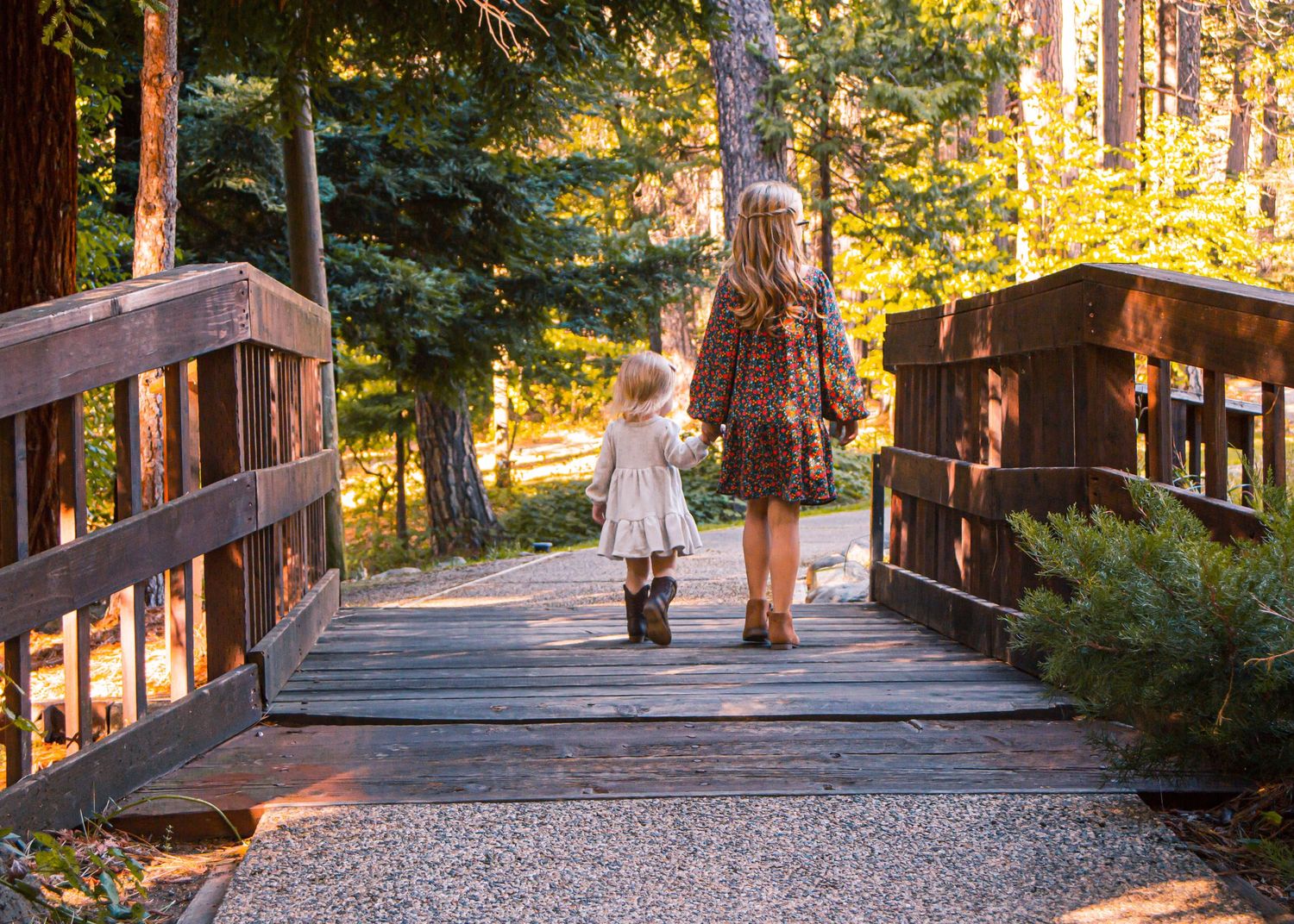 Big sister and little sister, holding hands, walking away from photographer across bridge, backs turned.
