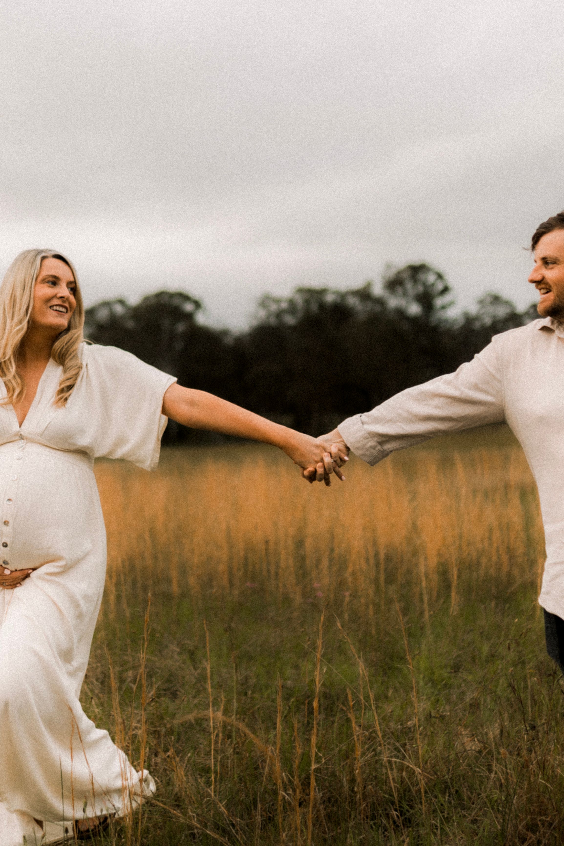 Expecting couple holds hands while walking through tall grass at sunset.