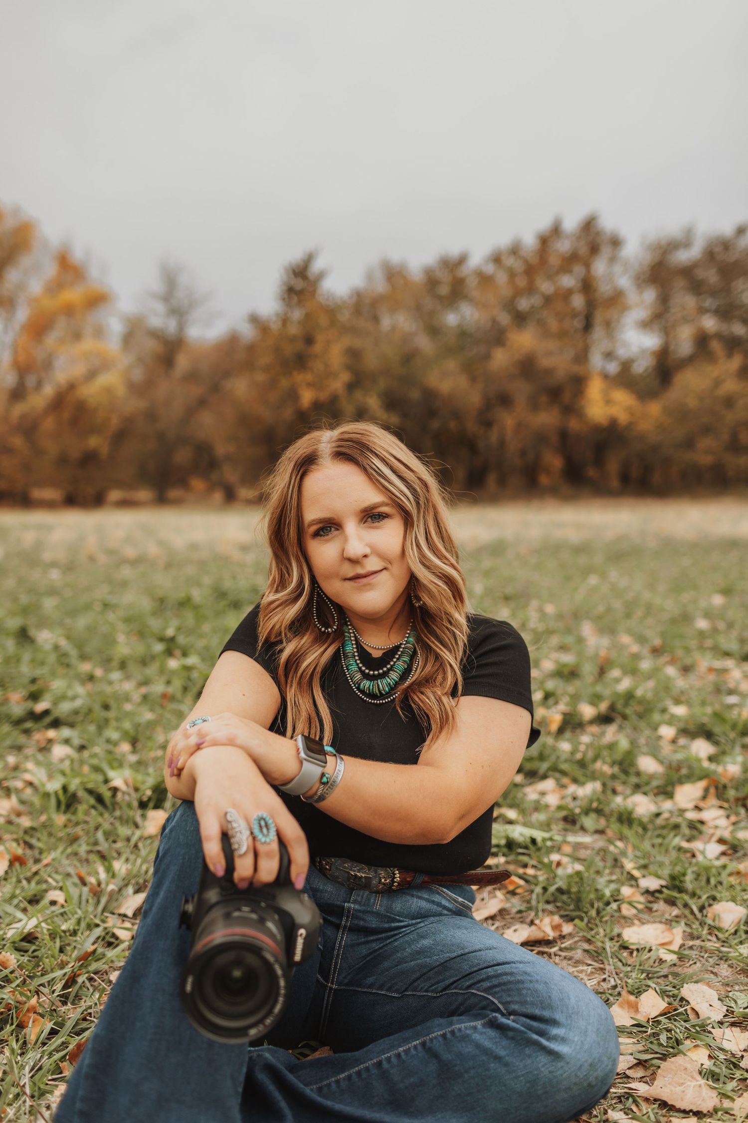 Professional photographer sitting in autumn field with camera wearing casual black top and jeans in golden light.