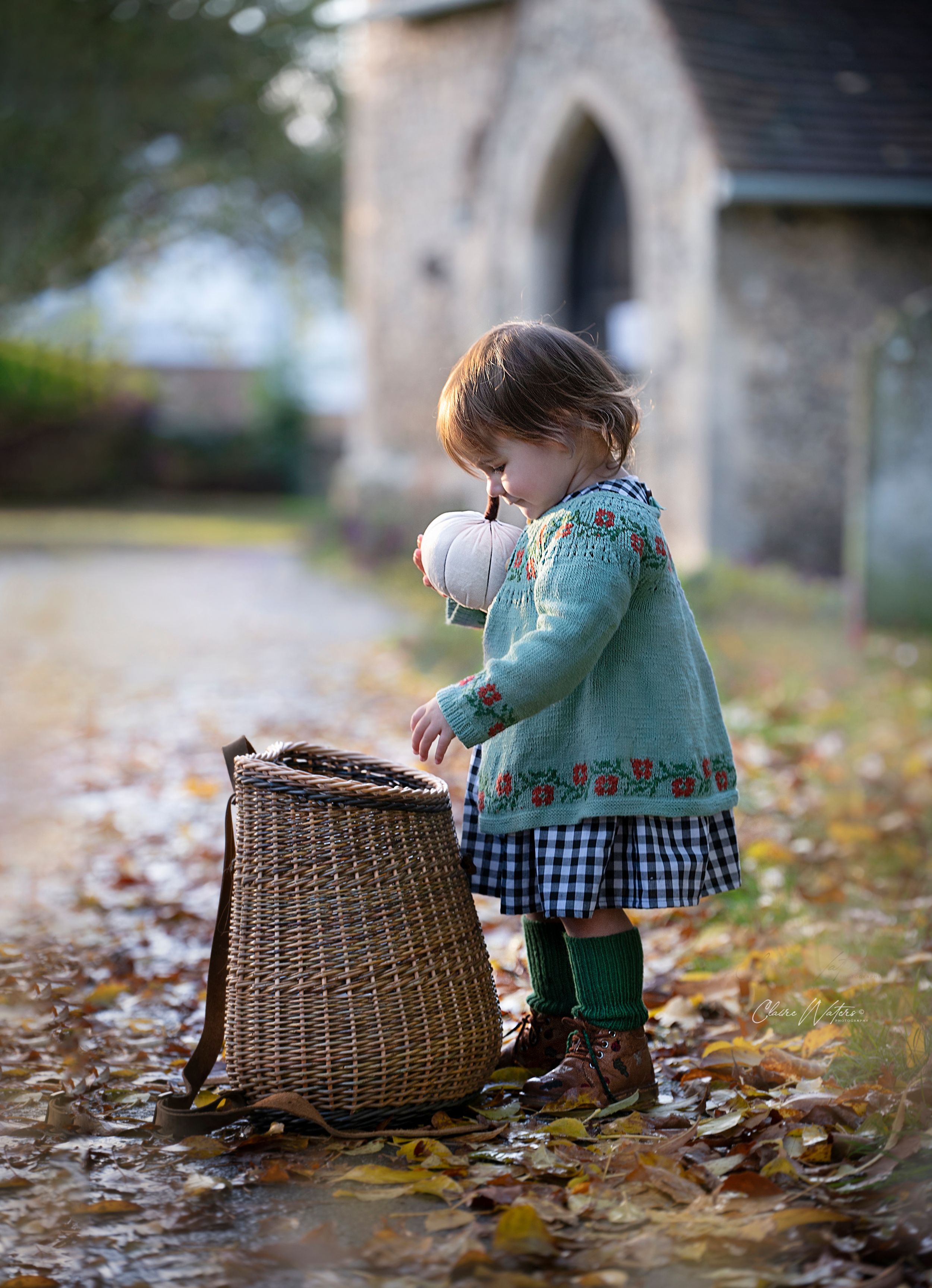 A small child in a teal sweater and boots explores a wicker basket surrounded by autumn leaves near a stone church entrance.