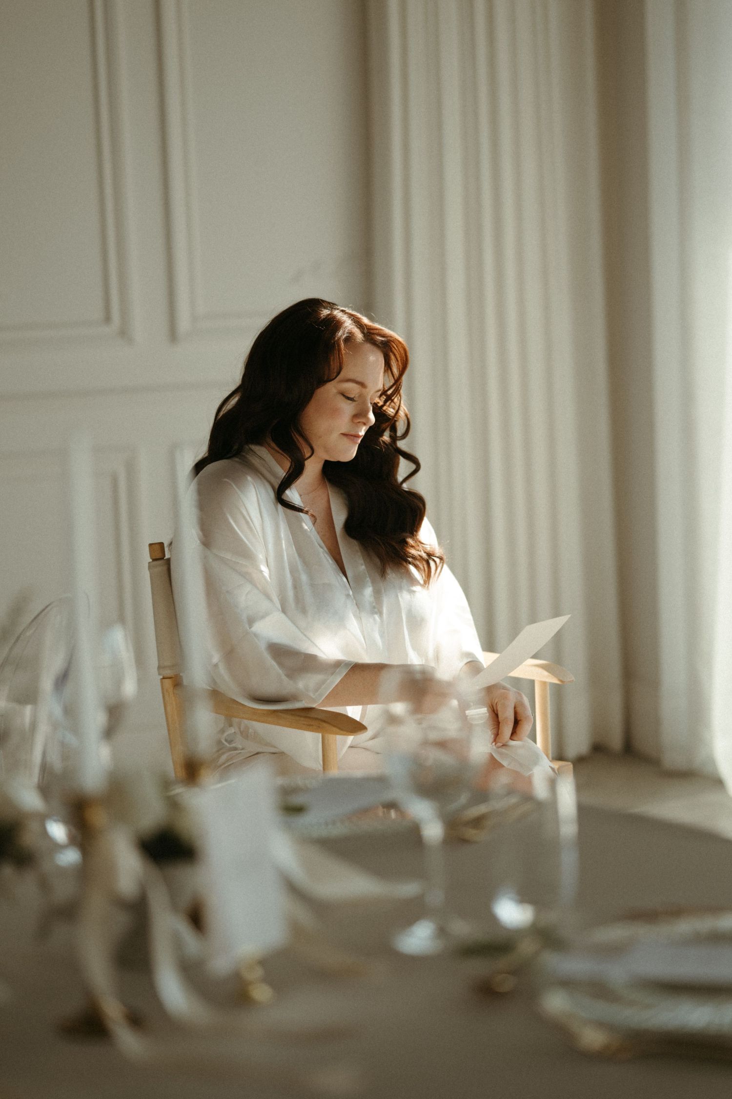 A woman in white sits at a table by a bright window in a serene indoor setting with soft natural lighting.