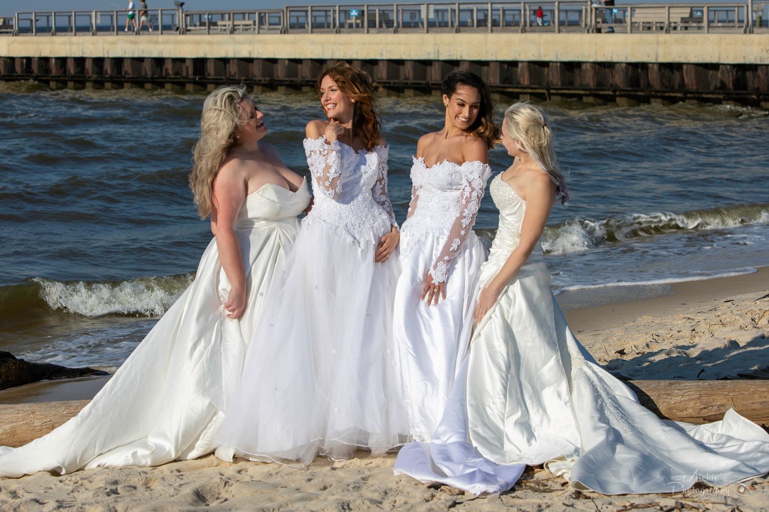 Four women in white wedding dresses posing together on beach near pier.