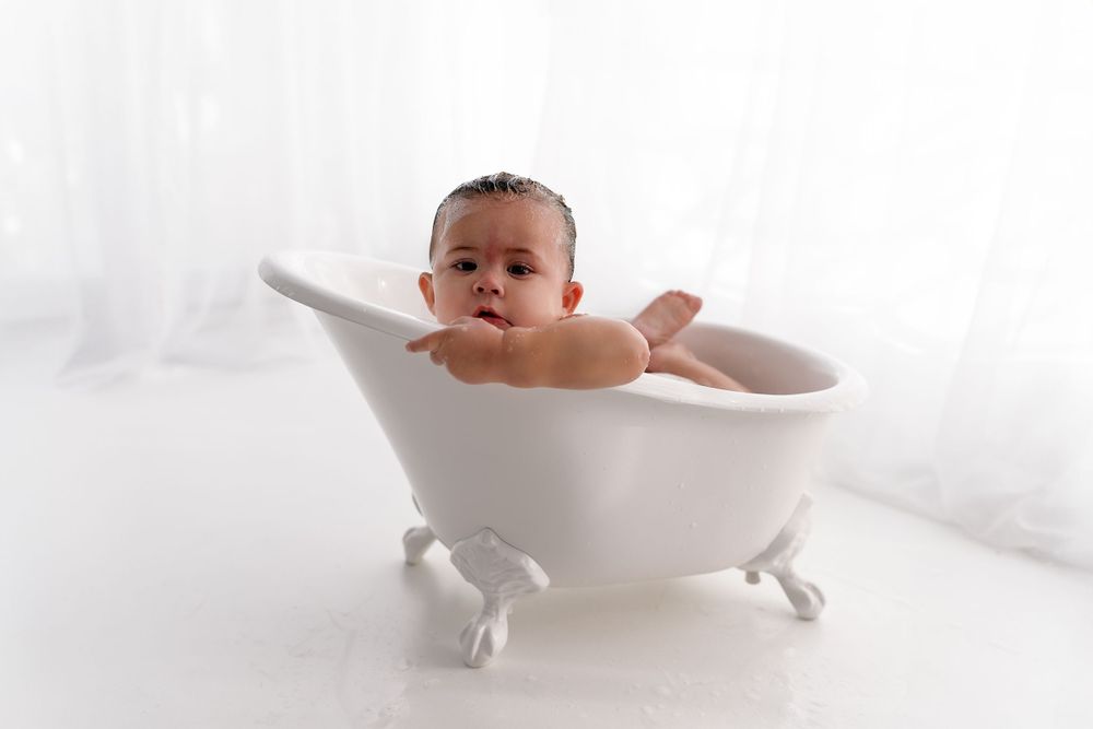 Baby peeking over edge of white clawfoot bathtub in studio setting.